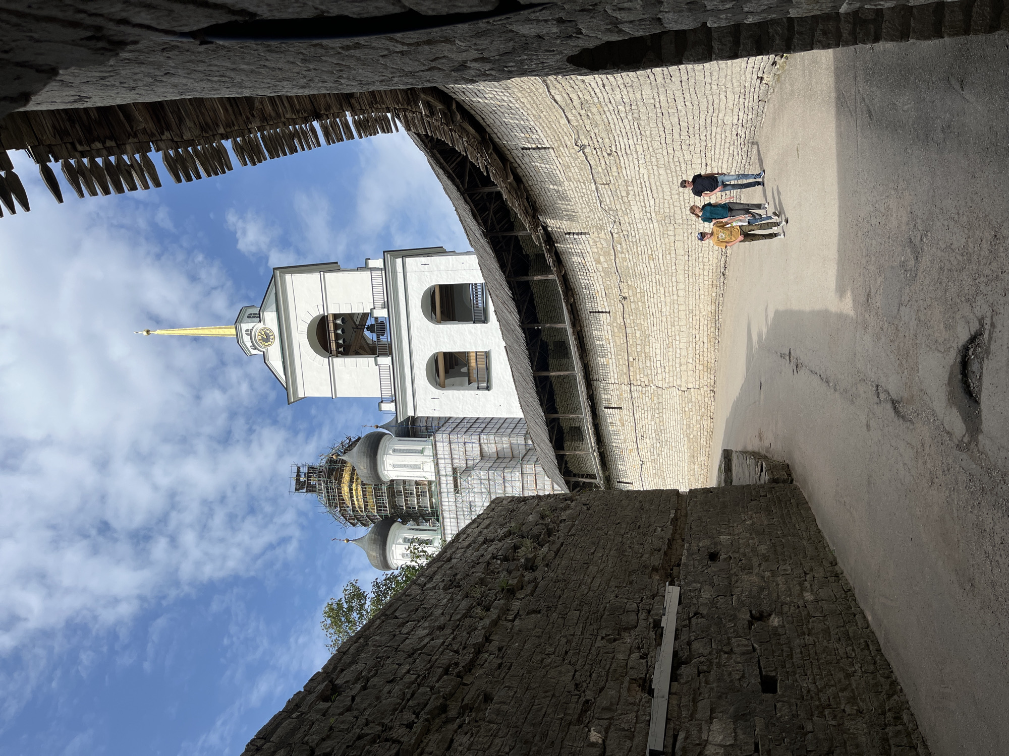 Three people, two adults and one child, walk along a paved path within the walls of the Pskov Kremlin in Pskov, Russia. The path is bordered by a thick, rough-hewn stone wall on the left and a lighter, more uniform brick wall topped with a wooden walkway on the right. In the background, a white bell tower with a golden spire stands tall, and behind it, a golden dome of a church is visible, partially covered by scaffolding, indicating ongoing restoration. The sky above is a mix of blue with scattered white clouds. The lighting suggests it is daytime, with sunlight casting shadows on the path.