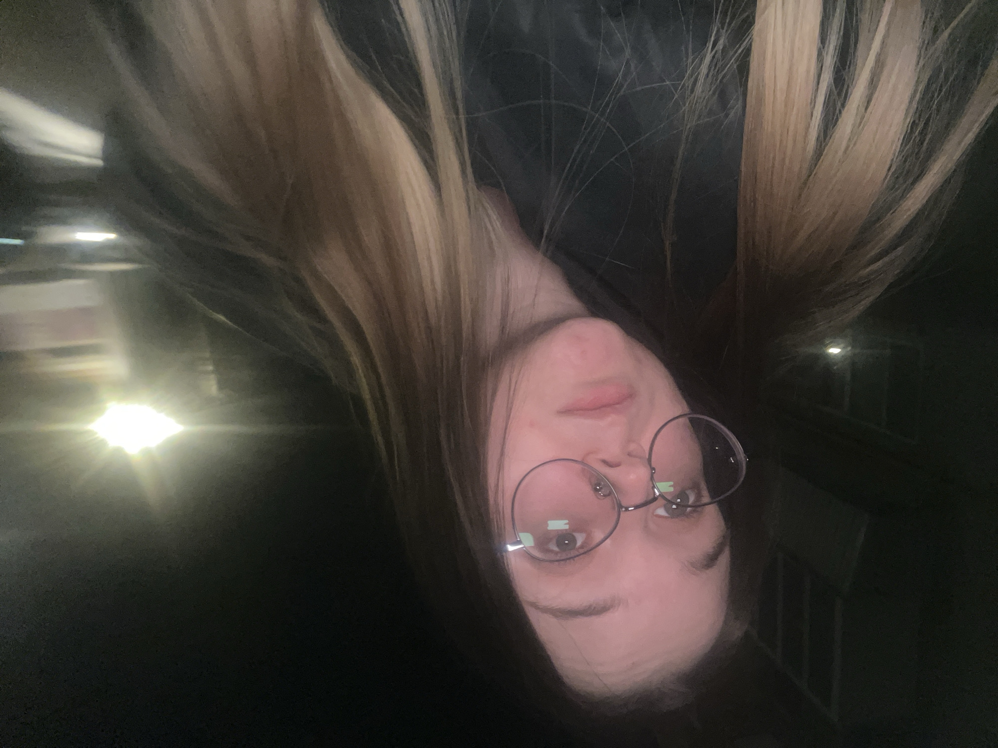 The scene is set in the city of Pushchino, Russia, at night. A female subject with light skin and long, two-toned hair (dark brown transitioning to blonde/light brown) occupies the majority of the frame, facing the camera directly. She wears round, silver-framed eyeglasses and a dark-colored jacket or coat. Her expression is neutral. The background is dark and blurred, indicative of nighttime conditions. A prominent, bright artificial light source with significant glare is visible in the upper right quadrant, consistent with an urban street or building light. Faint, diffuse lights are also present in the distant background, along with indistinct dark structures, possibly building facades or windows, faintly visible in the upper left background.