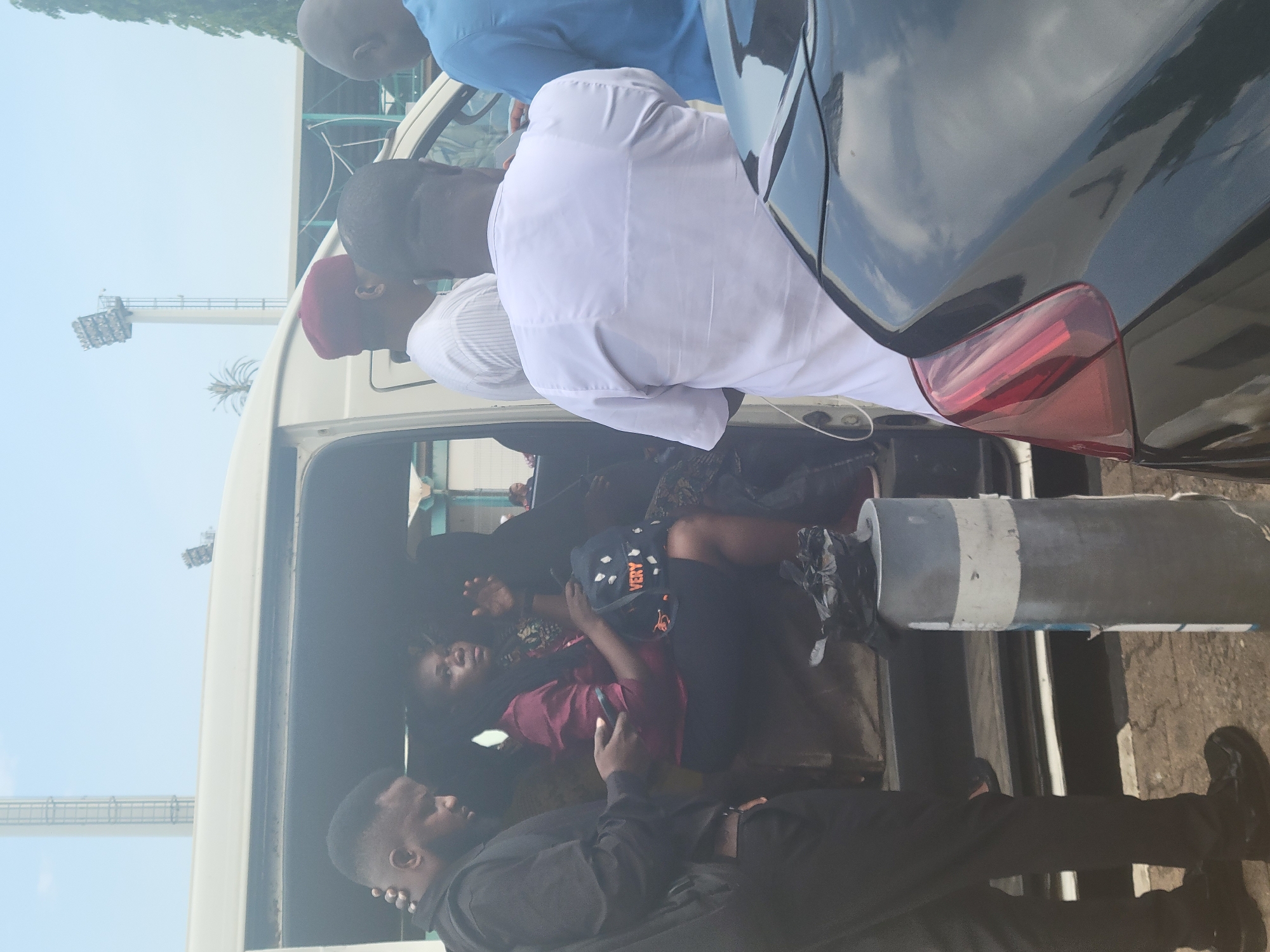 A daytime exterior shot shows several people gathered around the open side door of a white van, appearing to assist individuals inside. The people inside the van include a woman with dark hair wearing a dark cap with the word “VERY” written on it. The surrounding area appears to be an urban environment with buildings and a stadium visible in the background. A black sedan is parked close to the van on the right. The location is likely Abuja, Nigeria.