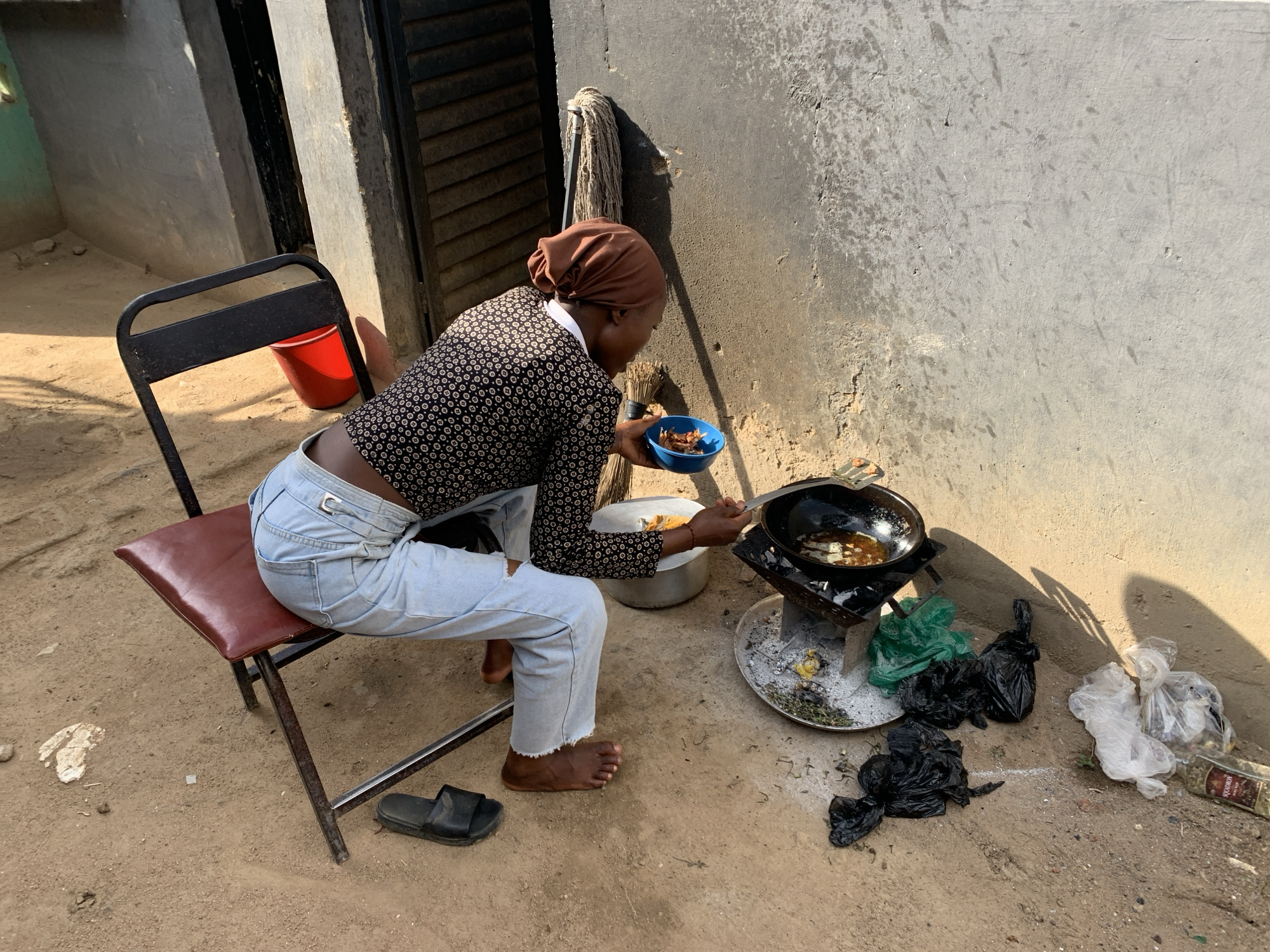 A person is cooking outdoors in Kabong, Nigeria. The individual, wearing a brown headscarf, a patterned long-sleeved shirt, and light blue jeans, is seated on a red chair. They are holding a small blue bowl in one hand while stirring food in a black frying pan placed on a metal stove. The stove is set on a large, flat, round metal tray. Several black and clear plastic bags are scattered on the dirt ground to the right of the stove. A pair of black sandals sits near the chair. A mop leans against a concrete wall behind the person. A red bucket is visible next to a doorway in the background. The ground is dirt. The setting appears to be in a residential area.