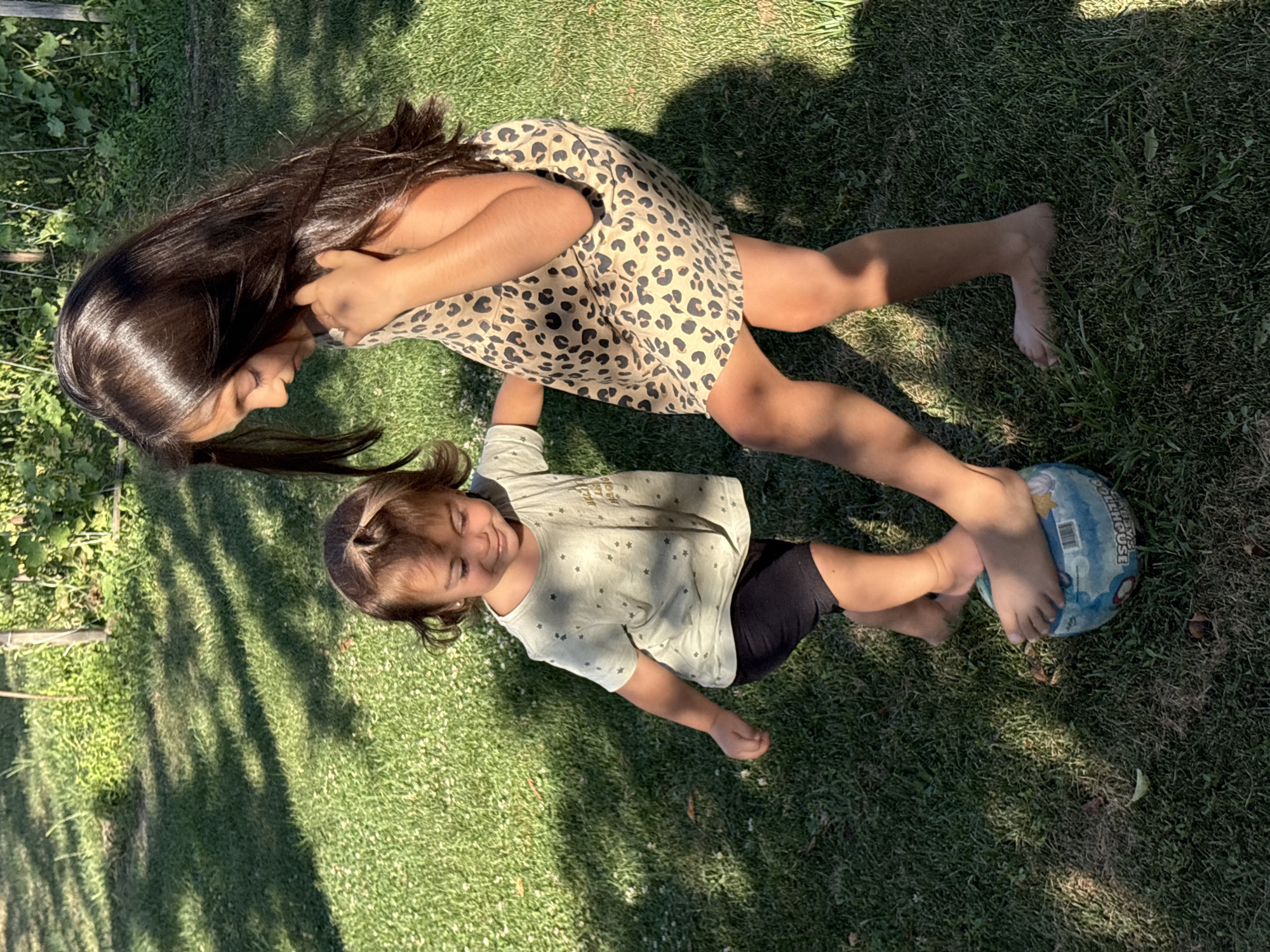 Two young girls are playing with a ball on a grassy lawn in a garden. The older girl, on the right, has long dark hair and is wearing a leopard-print dress. The younger girl, on the left, has dark hair and is wearing a light-colored t-shirt with small stars and black shorts. The older girl is standing with her foot on the ball, while the younger girl appears to be reaching for it. The background shows green grass, some foliage, and dappled sunlight and shadows. The image was taken in the city of Oreshak, Bulgaria.
