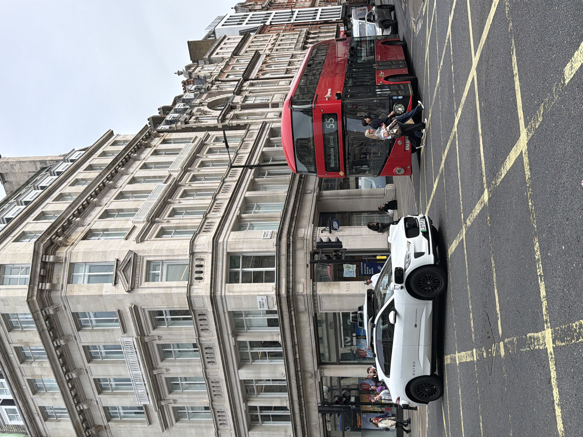 Here is a detailed description of the image:
The image captures a street scene in London, United Kingdom, showcasing a mix of urban elements. The street features a red double-decker bus with the number 55 heading towards Walthamstow Central. Passengers are seen both boarding and disembarking from the bus.
A white Waymo vehicle is parked to the side, indicating autonomous driving technology is being tested. There are several pedestrians in the image, some near the bus stop and others standing along the pavement.
The background prominently displays a large, multi-story building with classical architectural details. A street sign reading "Regent Street W1" confirms the location within London. The street is marked with yellow box junction lines, a traffic control measure to prevent gridlock. Other smaller buildings can be seen in the background, indicative of a densely built urban environment.
Signs on buildings are visible for Boots and other shops. The general environment suggests a busy commercial area within London.