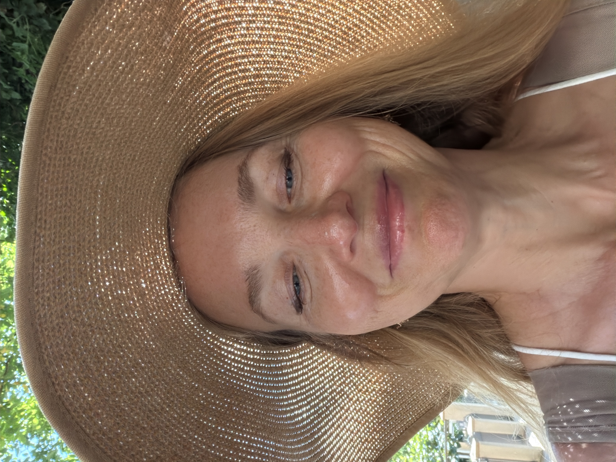 A woman wearing a wide-brimmed straw hat is taking a selfie outdoors. She has blonde hair and is smiling with her eyes partially closed. Her skin appears to have freckles and some sun exposure. She is wearing a light-colored top with thin straps. The background shows greenery, suggesting an outdoor setting, possibly a garden or park. The image does not provide any specific details about the location within the city of Budva, Montenegro.