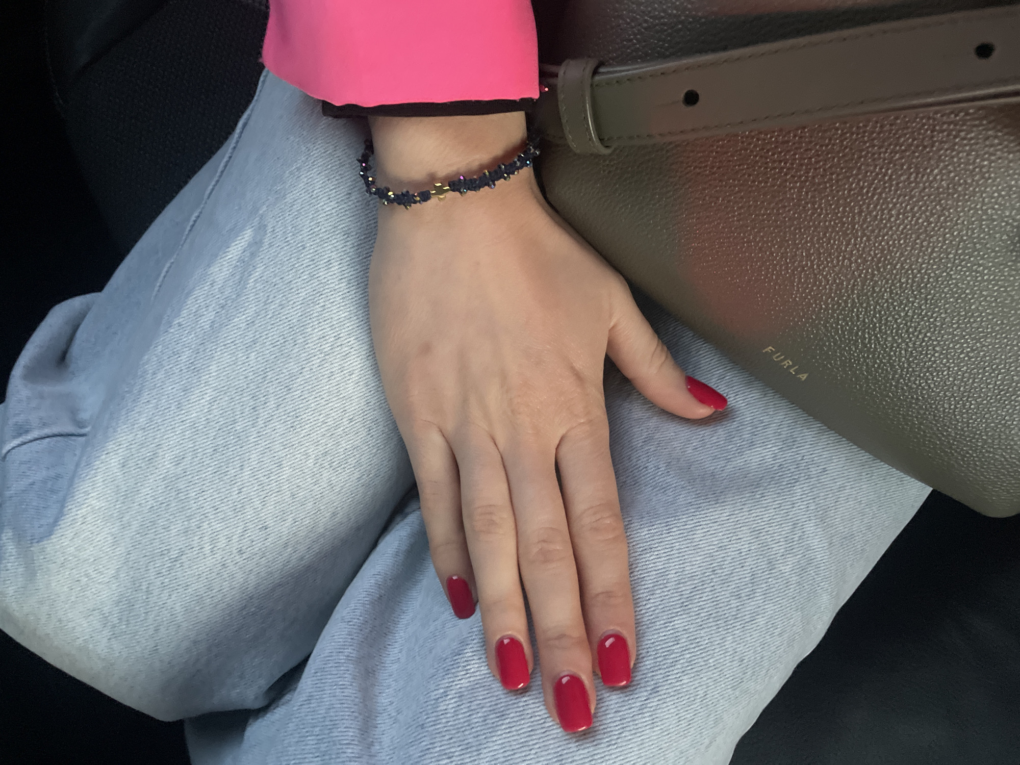 A photograph shows a person's hand resting on their leg. The person is wearing light-wash denim jeans and a bright pink top with a black trim. A gray leather bag with a strap is also visible. The person has red nail polish and is wearing a delicate bracelet with small dark stones and a gold accent. The bag has the word "FURLA" printed on it. The location is not discernible from the image, thus location is unknown.