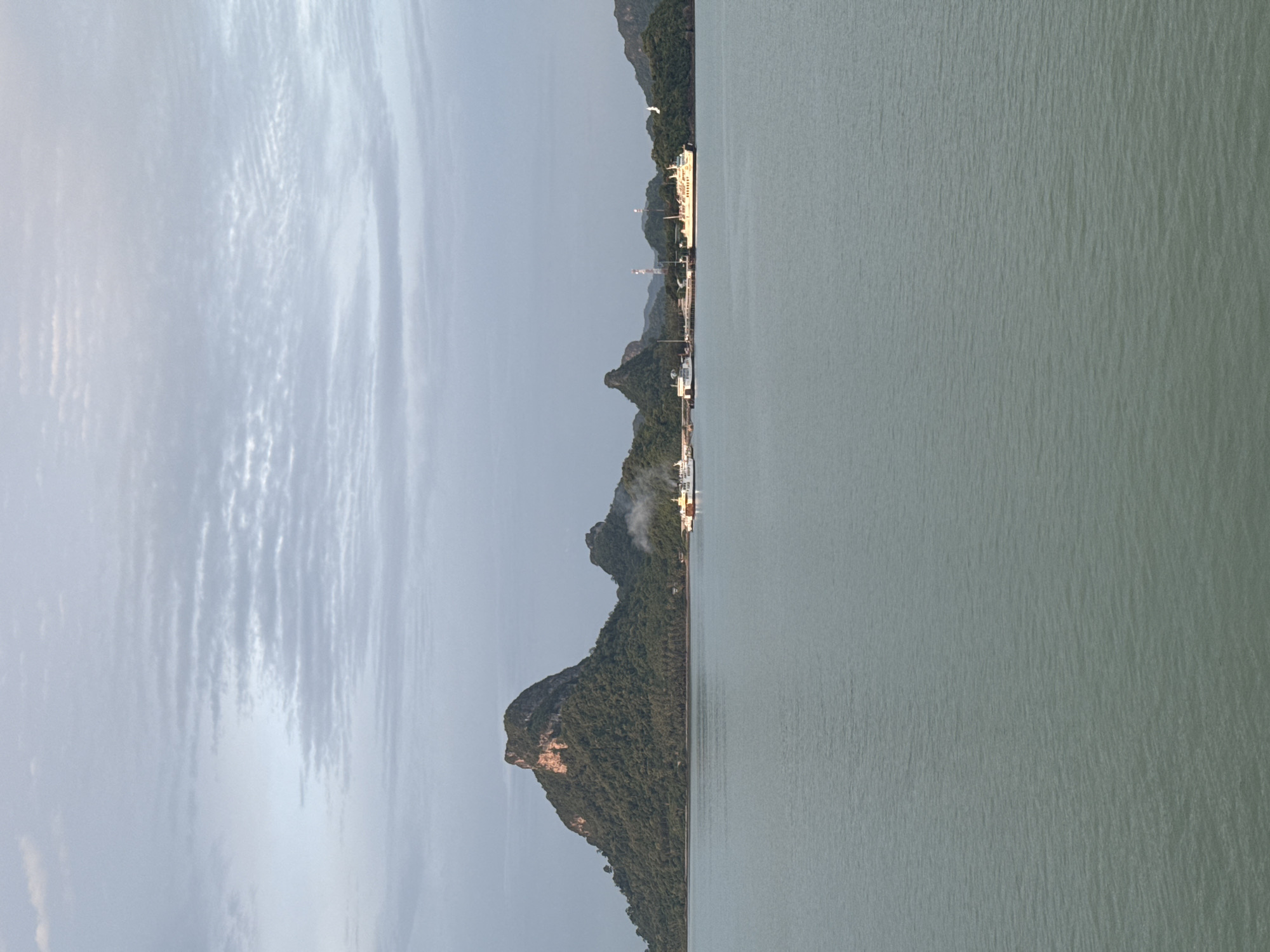 This image depicts a scenic view of the coastline near Don Sak, Thailand, at what appears to be either dawn or dusk, given the soft lighting and overcast sky. The foreground is dominated by a tranquil, slightly rippling sea, rendered in muted green and blue tones.
In the mid-ground, a lush, verdant island rises dramatically from the water, characterized by a prominent, craggy peak covered in dense vegetation. To the right of this main island, smaller, similarly forested islands stretch across the horizon. A small port or ferry terminal is visible along the coastline of the islands, with several vessels docked. These include at least two ferries, one of which is emitting a plume of white smoke, suggesting it is in operation or undergoing maintenance. The ferries appear to be white with yellow accents. Structures associated with the port, such as piers and possibly communication towers, are also discernible.
The sky above is a canvas of diffused light, with layered, textured clouds in shades of grey and pale blue, indicating a potentially humid or transitional weather condition. The overall atmosphere is serene and picturesque, highlighting the natural beauty of the Thai coast combined with maritime activity. There are no people visible in the frame.