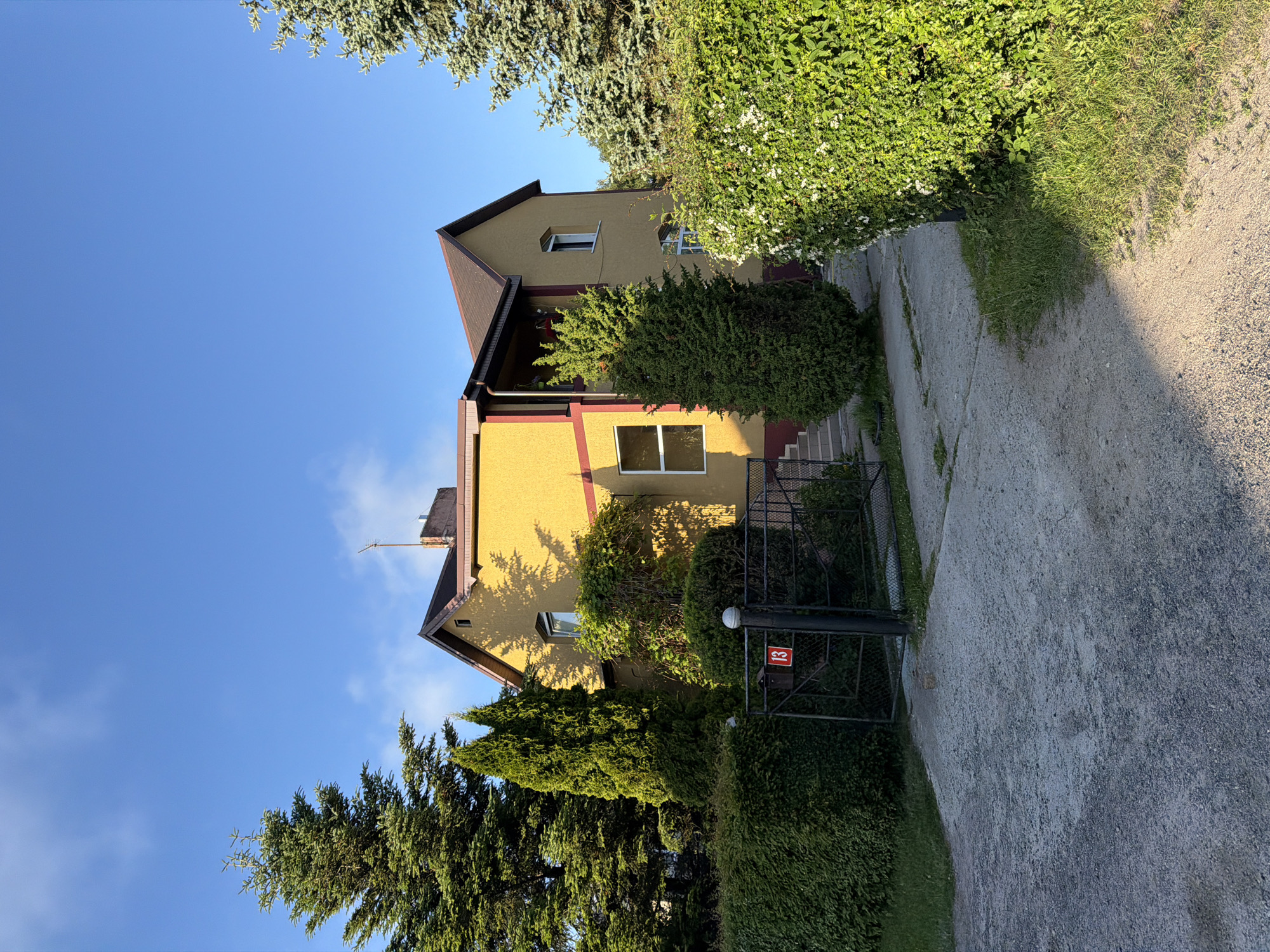 The image depicts a yellow house with a brown roof situated in Svetlogorsk, Russia. The house has several windows, some of which are visible from the exterior. The property is surrounded by lush green trees and hedges, creating a well-maintained garden setting. A gravel driveway leads up to the house, and a black metal gate with the number "13" displayed on it is visible at the entrance. The sky is a clear blue with a few wispy clouds. The lighting suggests it is daytime, with the sun casting shadows on the side of the house and the ground. The overall scene conveys a sense of suburban tranquility and domesticity.