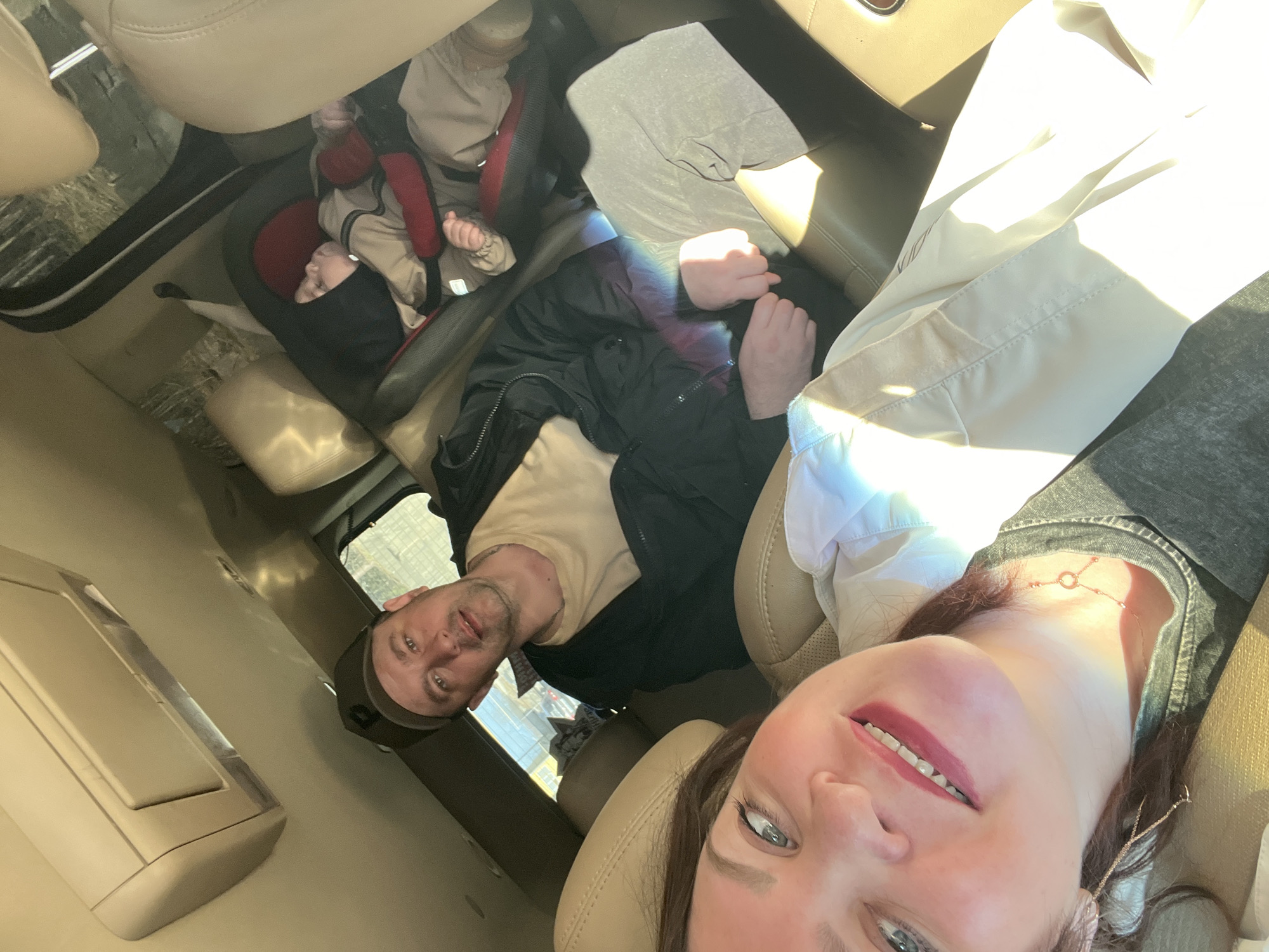 This media file captures a family inside a car, likely traveling, in Noviy, Russia. The image is taken as a selfie, rotated horizontally, giving a unique perspective of the vehicle's interior and its occupants.
In the foreground, a woman's face is prominent, looking directly into the camera with a subtle smile. She has light-colored eyes and is wearing red lipstick. Her neck is adorned with a delicate, gold-toned necklace featuring a circular pendant. She appears to be wearing a light-colored, possibly white, collared top over a darker garment, perhaps denim.
Behind the woman, a man is visible, also looking towards the camera. He wears a beige t-shirt, a black jacket, and a dark baseball cap. His expression is neutral. Further back, in the rear of the car, a baby is secured in a red and black car seat, appearing to be asleep or resting. The baby is dressed in a light-colored, possibly beige, outfit.
The setting is the interior of a car, characterized by beige or tan leather seats and an overhead console. Bright sunlight streams through the windows, casting strong highlights and shadows, indicating it is daytime and sunny. No specific weather conditions are discernible beyond the sunshine. The overall atmosphere suggests a casual family outing or journey.
There is no clearly legible text visible on any objects, clothing, or within the car's interior. The image primarily focuses on the family members and their immediate surroundings within the vehicle.