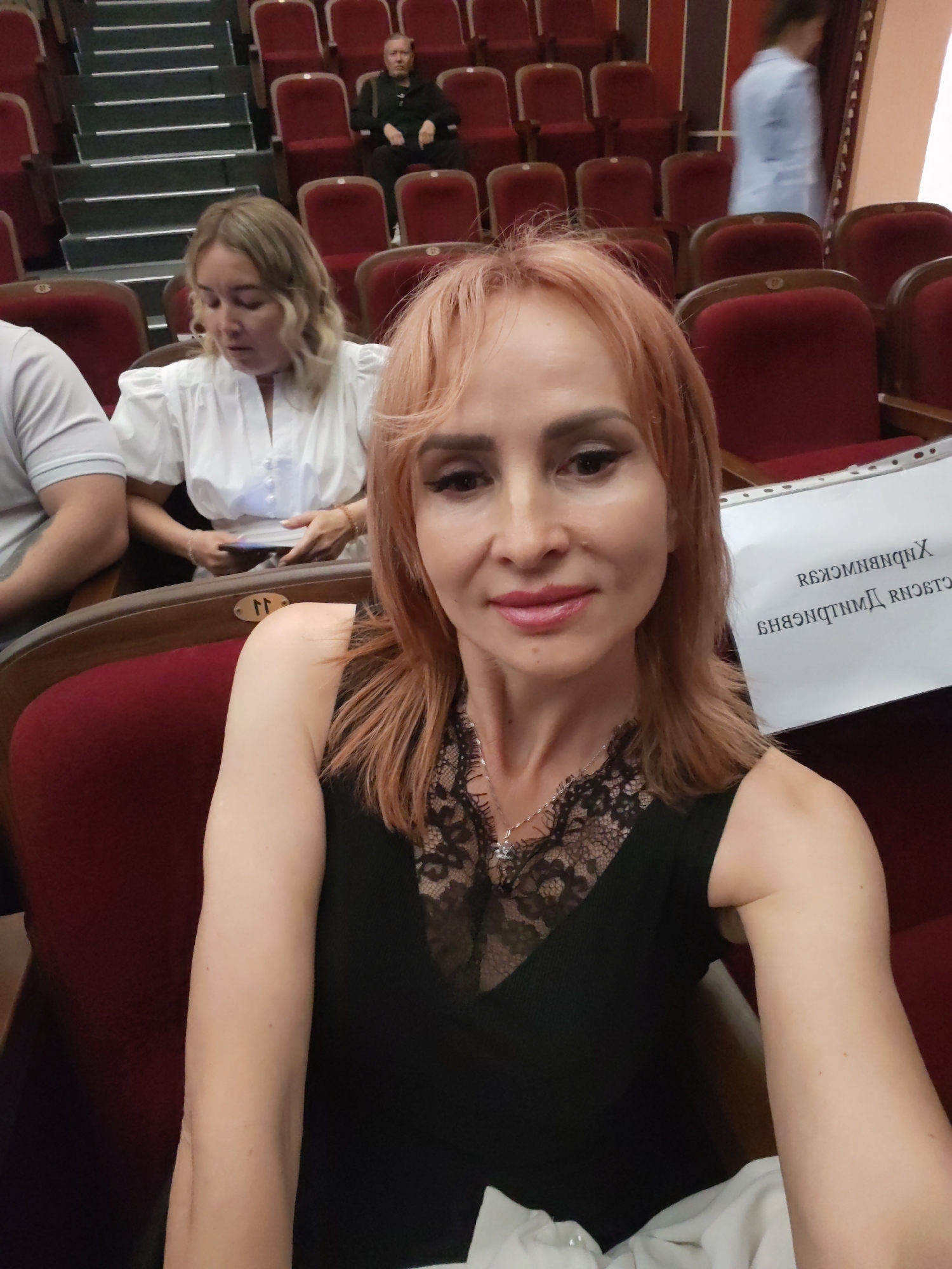 A woman with reddish-blonde hair is taking a selfie in an auditorium. She is wearing a black top with a lace V-neck and a silver necklace. Her right arm is resting on her lap, and her left arm is not visible. In the background, there are rows of red seats. To her left, a woman with blonde hair is sitting in a white top and holding a brochure. Further in the background, a man is seated in the upper rows. To the right of the selfie-taker, a piece of paper with text in a foreign language is propped up. The scene appears to be in the city of Chaikovsky, Russia, likely at a theater or concert hall.