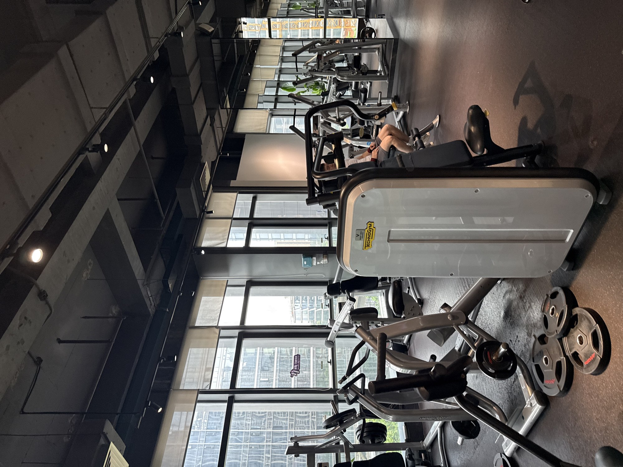 The image depicts the interior of a modern gym located in the Haizhu District, China. The gym features a dark ceiling with exposed beams and lighting tracks, and large windows overlooking a cityscape with tall buildings. The floor is dark rubber matting, typical for gyms.
Several pieces of weightlifting equipment are visible, including a Technogym machine in the foreground. Other machines, characterized by their metallic frames and padding, are arranged throughout the space. Weight plates are scattered on the floor near some of the equipment.
In the mid-ground, a young man is seated on a weight machine, wearing a white t-shirt and grey shorts. He appears to be engaged in a workout. To the right, a large mirror reflects more gym equipment and the cityscape outside. Natural light filters through the blinds covering the large windows. The overall impression is a well-equipped, contemporary fitness facility.