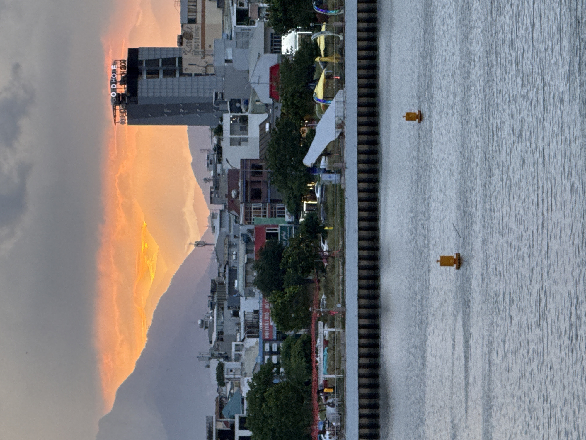 This is a photograph of the cityscape of Da Nang, Vietnam, taken during sunset. The image features a body of water in the foreground with a buoy floating on its surface. In the middle ground, there is a waterfront promenade with buildings and trees. The buildings are a mix of residential and commercial structures, with some featuring illuminated signs. The background shows mountains, a tall building with the "OD HOTEL" sign, and a dramatic sunset sky with orange and gray clouds. The overall impression is one of a coastal city at dusk.