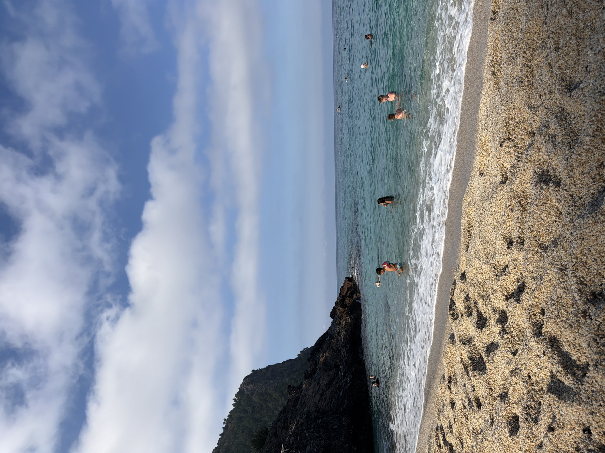 A vertical, eye-level medium shot shows a rocky cliff face on the left, a calm turquoise sea with several people swimming, and a sandy beach in the foreground with many footprints. The sky is blue with scattered white clouds. The cliff face is dark and rocky, with trees and vegetation growing on top. The sea is clear and shallow near the shore, with white waves breaking on the sand. Several people are visible in the water, some swimming, others standing. In the foreground, the sand is light brown and coarse, with deep footprints forming patterns. The lighting suggests it is daytime.