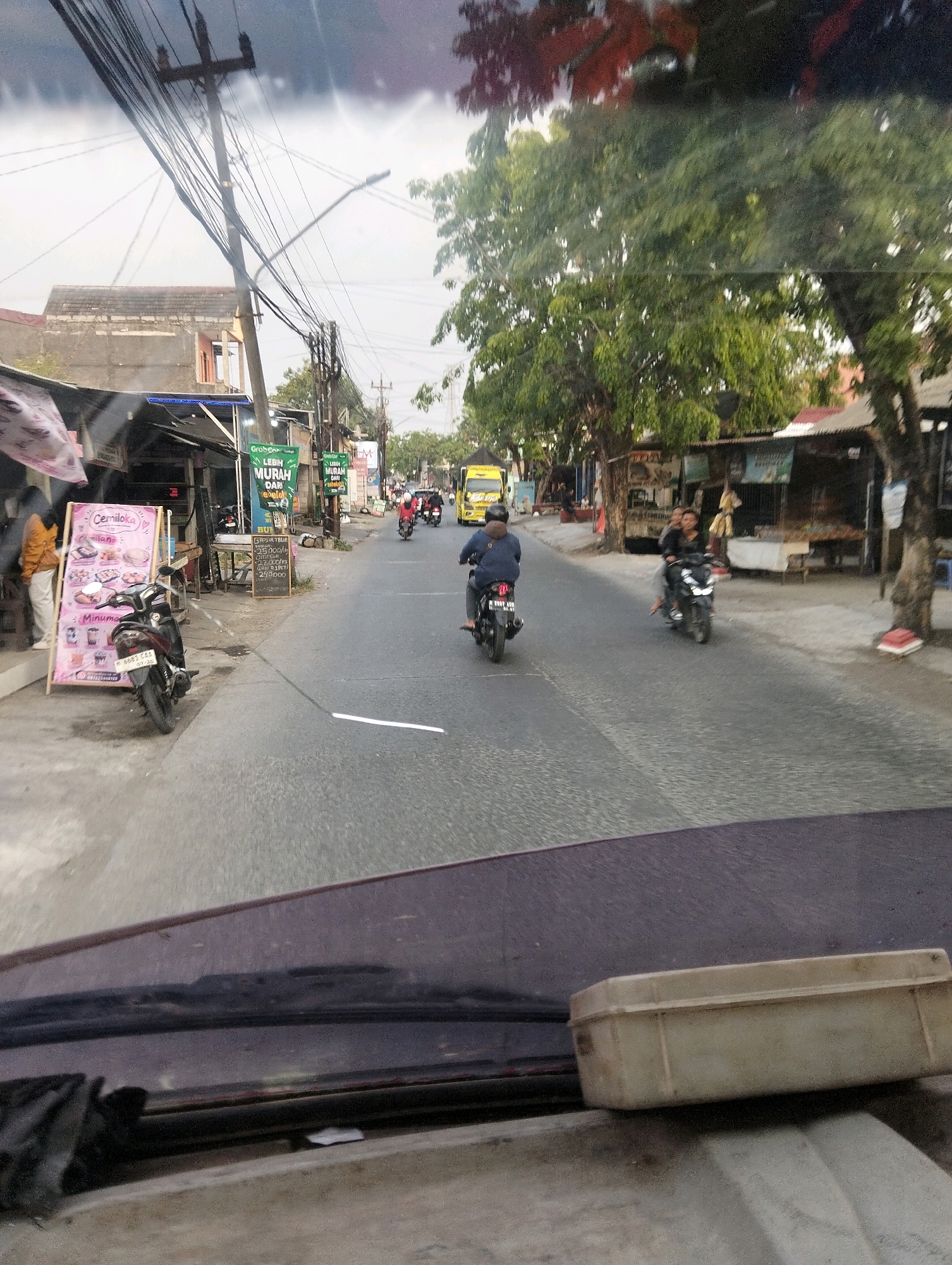 The image depicts a street scene in Semarang, Indonesia, viewed from the perspective of a vehicle's interior. The road is paved and appears to be a two-lane street with some vehicles and pedestrians present. On the left side of the road, there are several small shops or stalls. One stall prominently displays a pink advertisement board with the text "Cemiloka" and images of food items. Next to this stall is a parked motorcycle. Further down the street, there are more shops with awnings and signs, including one with green text that reads "GRABFood LEBIH MURAH DARI". A utility pole with numerous electrical wires stands on the left side of the street, close to the shops.
On the road, several motorcycles are visible. In the foreground, a person wearing a blue jacket is riding a motorcycle, facing away from the camera. Further ahead, a yellow truck is driving in the same direction. Two people are riding on another motorcycle, with the driver wearing a dark helmet and the passenger in front of them. On the right side of the road, there are more shops and trees lining the street. A few other motorcycles are also seen in the distance, moving along the road.
The foreground of the image is dominated by the interior of a vehicle, likely a car, with a portion of the dashboard and windshield visible. A beige rectangular object, possibly a container or a box, rests on the dashboard. The sky is overcast, suggesting a cloudy day. The overall impression is that of a typical urban street scene in Indonesia, with everyday activities taking place.