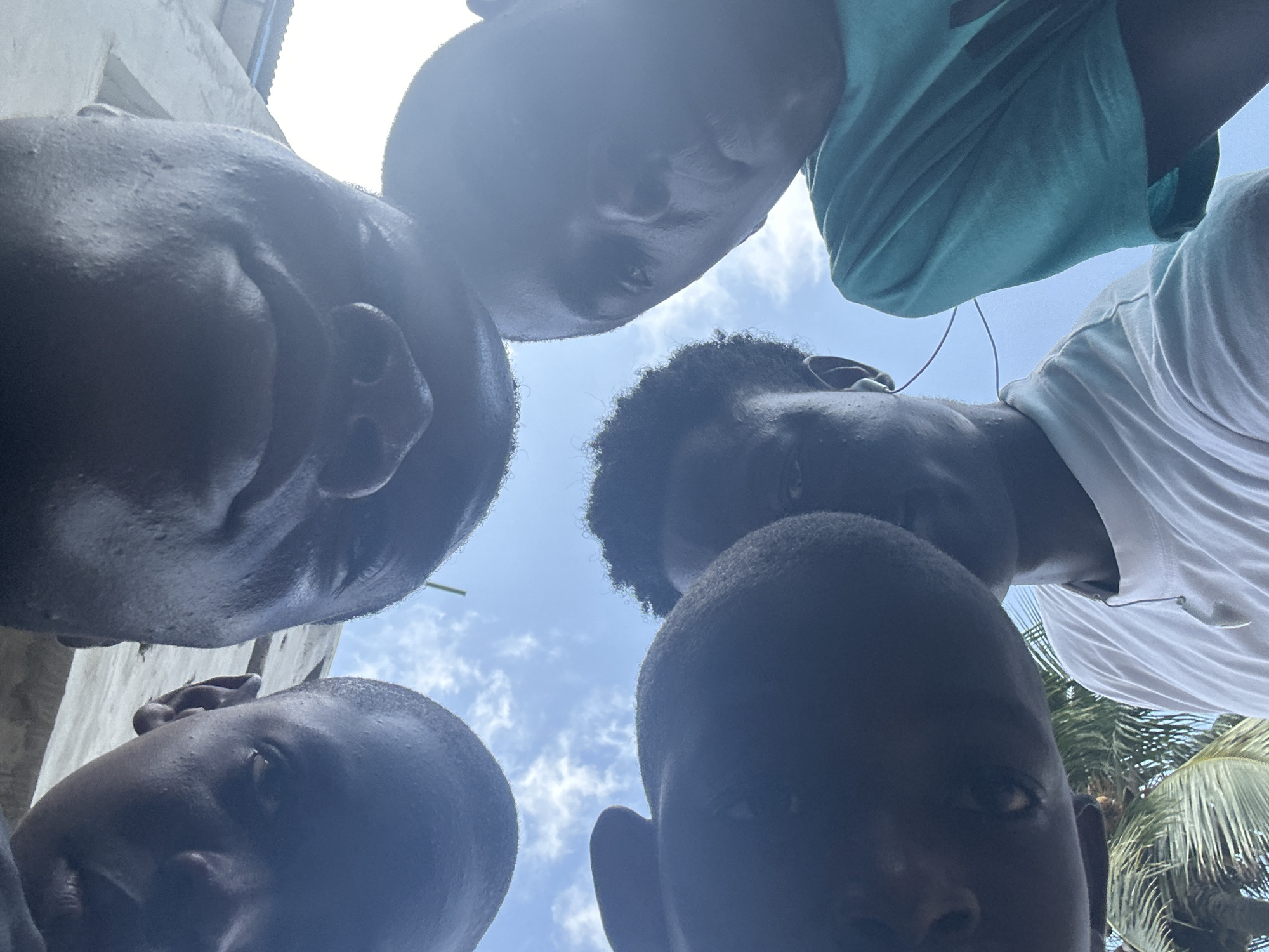 This low-angle photograph captures five young Black individuals looking down directly into the camera, creating a unique perspective as if the viewer is on the ground. The subjects are positioned in a semi-circle, with their heads and shoulders filling most of the frame, silhouetted against a bright sky.
Their skin appears dark, with some facial features discernible despite the challenging upward lighting. Their hair is short or curly. Two individuals are wearing t-shirts: one on the top right in a light blue or teal shirt, and another on the mid-right in a white t-shirt.
The scene is set outdoors under a bright blue sky with scattered white clouds, indicating clear, sunny weather during daytime, likely midday or early afternoon given the intense overhead light. In the background, glimpses of a light-colored building or wall are visible on the left side, and lush green palm tree fronds can be seen on the right, hinting at a tropical or subtropical environment. This aligns with the provided context that the location is Badagry, Nigeria.
The activity appears to be the group gathering and looking down with a sense of curiosity or engagement towards the camera. No visible text is present in the image.