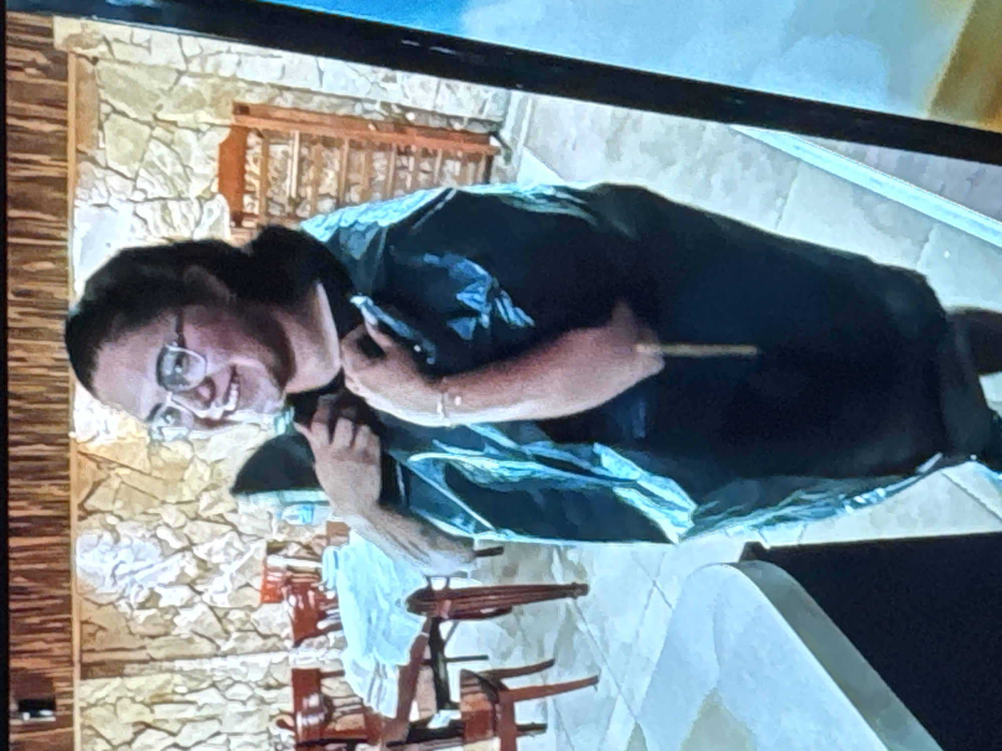 The image shows a young woman wearing glasses and a large black trash bag over her body. She is smiling and looking to her left, with her hands near the opening of the bag. The background appears to be an indoor setting with stone walls and wooden furniture, including tables and chairs. A wooden shelf unit is visible in the background. The lighting is somewhat dim. This scene is not located in the city of Sredets, Bulgaria, as there are no visual cues to suggest this location.