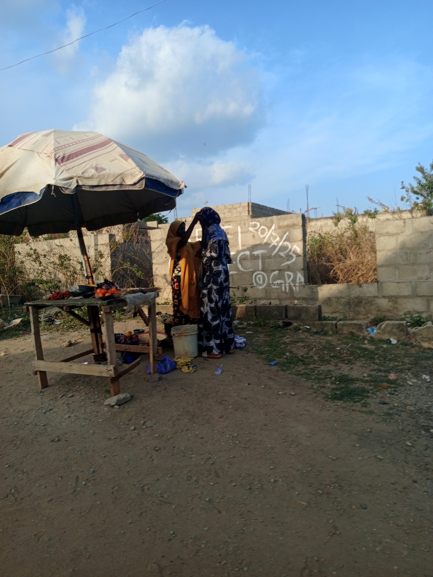 Two women in traditional African dress stand near a makeshift fruit stand in Keffi, Nigeria. The stand, shaded by a faded umbrella, displays tomatoes, oranges, and other produce. The women appear to be interacting; one is touching the head covering of the other. A concrete wall with graffiti is visible in the background. The ground is dusty and strewn with litter. The sky is blue with scattered clouds. The location is a roadside.