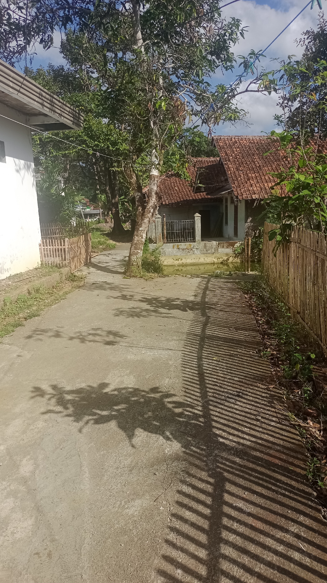 The image depicts a concrete pathway in Tonjong, Indonesia, leading towards a traditional house with a red tile roof. The pathway is flanked on the right by a bamboo fence, and on the left by a white building with a gray roof overhang and a wooden fence. Lush green trees and foliage surround the scene, casting dappled shadows on the pathway. The sky above is blue with scattered clouds. The overall impression is of a rural residential area.