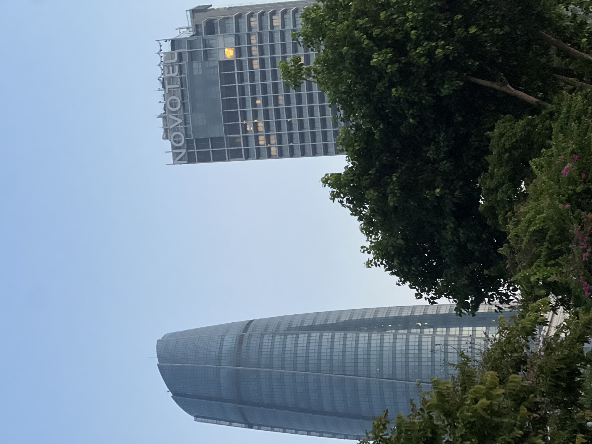 The image shows two modern skyscrapers against a clear blue sky. The skyscraper on the left is taller and has a curved, glass facade. The skyscraper on the right is shorter and features the "NOVOTEL" logo prominently displayed at its top. The lower portion of the image is filled with lush green trees and foliage, partially obscuring the bases of the buildings. Some windows in the Novotel building are illuminated, suggesting it is evening or nighttime. This scene depicts the cityscape of Đà Nẵng, Vietnam, likely near the Dragon Bridge area, a prominent landmark known for its modern architecture and riverside developments.
