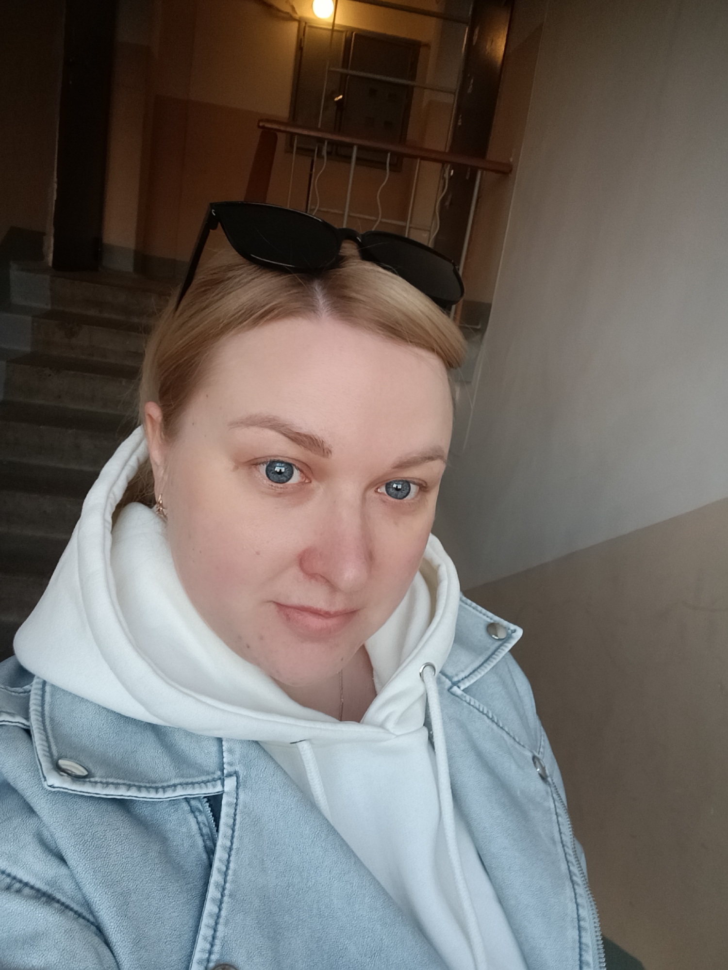 A selfie shows a blonde woman with blue eyes wearing a white hoodie under a light blue denim jacket. Sunglasses rest on her head. She is inside, possibly in a stairwell of an apartment building, evidenced by the stairs, railing, and lighting visible in the background. The location is Chaikovsky, Russia, based on the description provided.