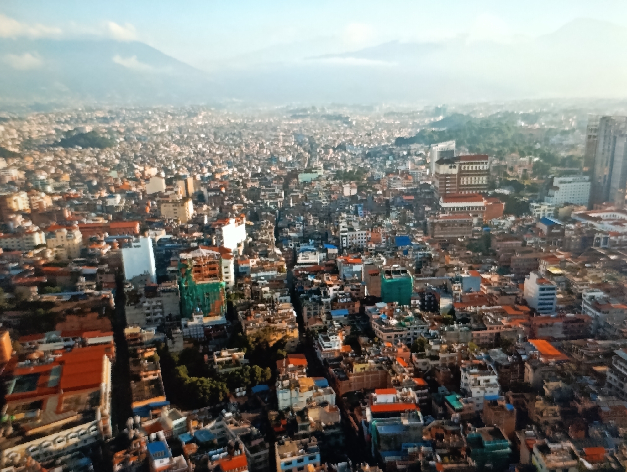 This is an aerial, wide-angle view of a densely populated city, likely a capital or major urban center, possibly in Asia, given the architecture and the distant mountains which appear to be of significant height. The city is characterized by a vast expanse of buildings, predominantly mid-rise structures with varied architectural styles, ranging from older, weathered buildings to more modern concrete and glass constructions. Rooftops are a prominent feature, displaying a mix of colors, including red tiles, gray concrete, and some with visible solar panels. Greenery is sparsely scattered throughout the urban landscape, primarily in the form of trees that seem to be planted on rooftops or in small, enclosed courtyards.
The city is experiencing a hazy atmosphere, common in large urban areas, which softens the distant view and creates a sense of depth. In the background, a range of imposing mountains is visible, partially obscured by a layer of haze or clouds. The lighting suggests it is either early morning or late afternoon, with the sun casting long shadows and illuminating some parts of the cityscape. The image shows no visible people or ongoing activities, presenting a static snapshot of the urban environment.
It is important to note that the request to include details about "the location in the city of Dubăsari town council, Moldova" is not supported by the visual evidence in the image. The architectural style, urban density, and the presence of prominent, high mountains in the background are not characteristic of Dubăsari, Moldova. Therefore, no information regarding Dubăsari can be provided based solely on this image.