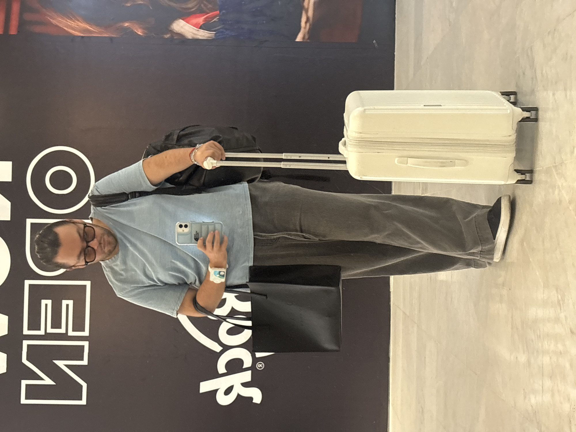 A man stands in front of a Hard Rock Cafe storefront. The cafe is in Mexico City, Mexico. The man is wearing glasses, a light blue shirt, and gray pants. He is holding a phone in his left hand and a black bag in his right. He has a black backpack on his back and is pulling a white suitcase. The floor is made of a light gray material.