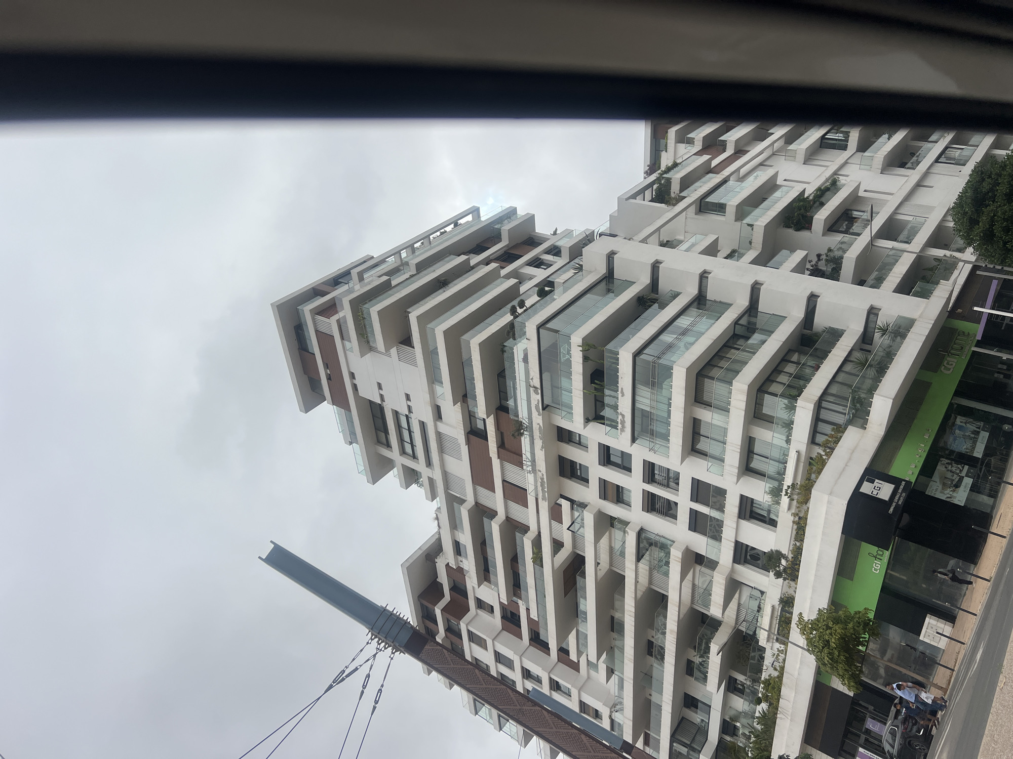 This image captures a modern, multi-story building from a low-angle perspective, likely from a vehicle or street level, under an overcast sky. The building, oriented vertically in the shot, features a distinctive architectural design with a white facade, numerous staggered balconies, and terraces. Many balconies are equipped with glass railings and hold various potted plants and greenery. Darker brown or wooden accents are visible between sections of the white structure.
At the ground level, the building houses commercial spaces with large glass windows. Prominently visible on a green and black shopfront are the words "CGI home," "CGI," "COMPAGNIE GENERALE IMMOBILIERE," and "CGI.MA," suggesting a real estate or construction company.
On the street below, a paved sidewalk is visible, with a dark SUV parked near the curb. Two individuals are seen walking on the sidewalk. A small tree is planted alongside the road. Extending into the frame from the lower left is a substantial grey structural element, resembling an I-beam, with several suspension cables attached, possibly part of a bridge or overhead infrastructure. The overall scene depicts a bustling urban environment during daytime under cloudy weather conditions.