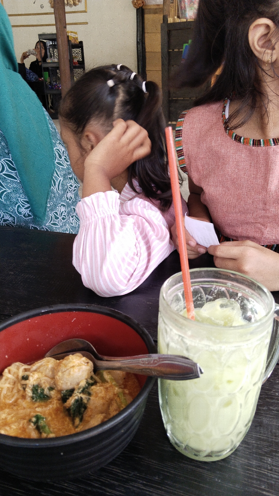 In a casual indoor setting, possibly a restaurant in Margatunggal, Indonesia, two individuals are partially visible. A young girl, on the left, is seen from the shoulders up, with her head turned away and her hand raised to her ear, wearing a striped pink and white long-sleeved shirt with ruffled cuffs. To her right, a person, likely an older child or adult, is seen from the chest up, wearing a pink top with a decorative, striped neckline, and their hands are visible holding a white piece of paper. In the foreground, a dark bowl contains a meal of noodles and what appears to be chicken in a creamy orange sauce, with a spoon resting inside. To the right of the bowl is a clear glass mug filled with a pale green beverage, ice, and a red straw. The background is blurred, showing shelves with items, suggesting a commercial establishment. The overall lighting is ambient, contributing to a relaxed atmosphere.