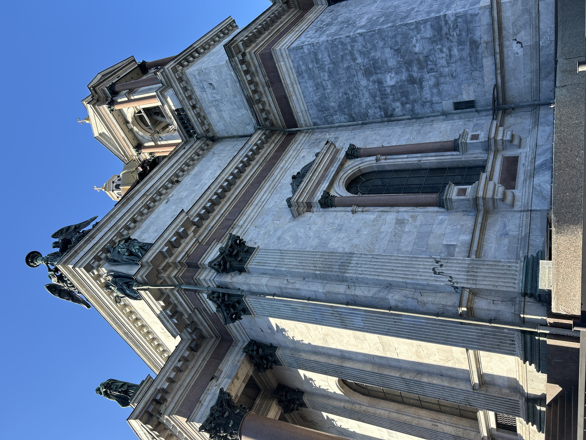 The image shows a low-angle view of the exterior of a large, ornate building under a clear blue sky. The building appears to be made of light-colored stone, possibly marble or granite, and features classical architectural details, including columns, cornices, and decorative moldings. The columns are topped with elaborate capitals, and there are statues and decorative elements near the top of the building. A few windows can be seen, framed by stone columns and decorative surrounds. The scene is well-lit, casting shadows on the building's facade. Given the architecture, it is likely a cathedral or other significant religious structure. Without additional context, the location in the city is unknown.