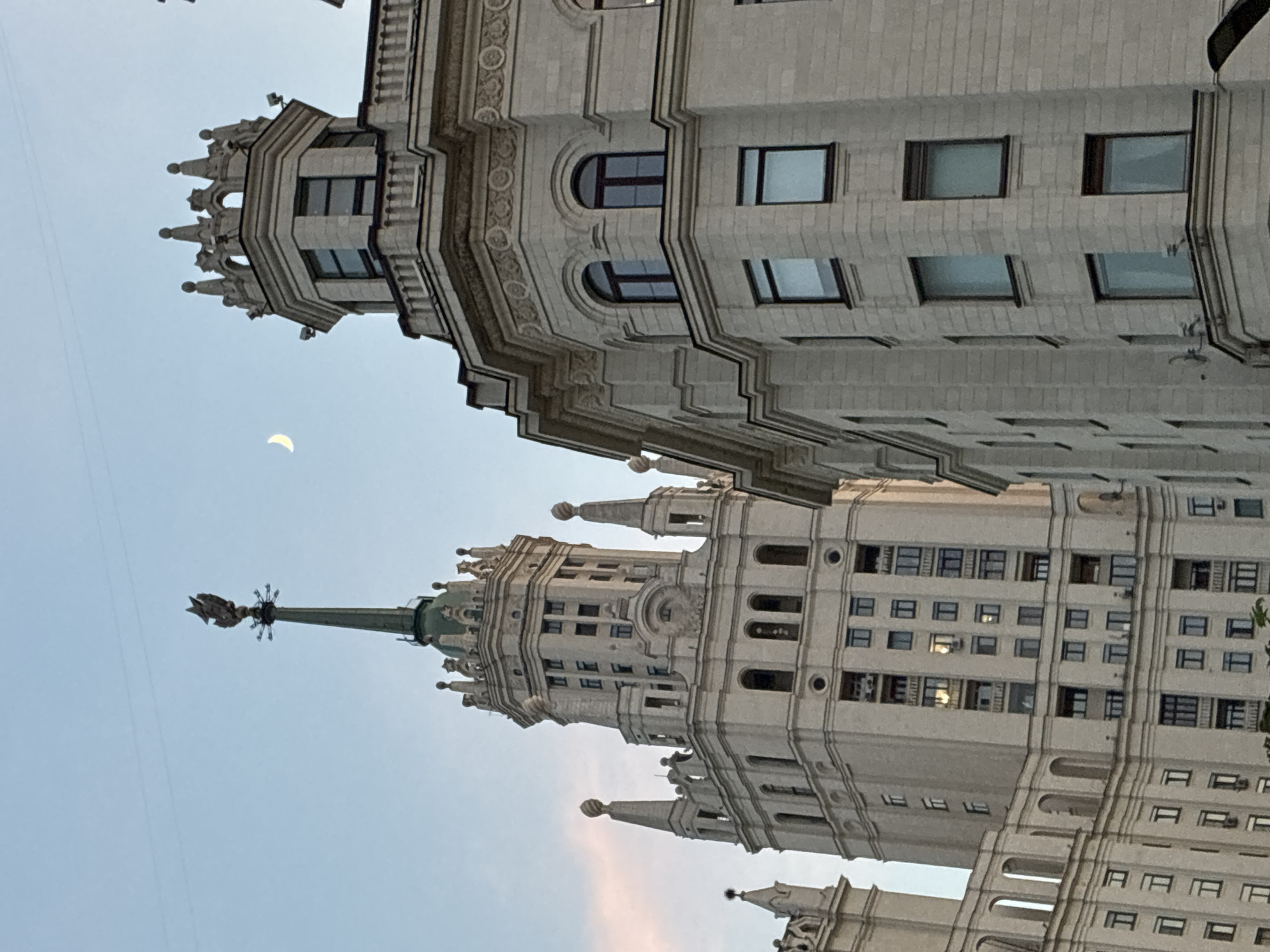 The image shows a low-angle view of the Kotelnicheskaya Embankment Building in Moscow, Russia, at dusk. The building's ornate, Stalinist architecture is prominently displayed, including its spires, decorative elements, and numerous windows. A crescent moon is visible in the sky. The sky is light blue with a hint of pink near the horizon. The perspective is slightly tilted.