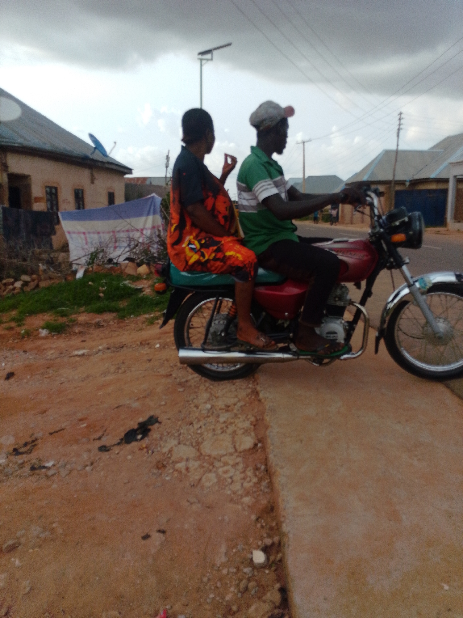 In Gombe, Nigeria, two individuals ride on a red motorcycle. The person in the front, a man wearing a green and white striped shirt and a grey cap, operates the motorcycle. The person behind him, a woman in a vibrant orange and black patterned dress, is seated with her legs visible. The motorcycle is on a dirt road next to a paved pathway. In the background, there are buildings with corrugated metal roofs, a satellite dish on one of the roofs, a street light, and utility poles with overhead wires. The sky is overcast with grey clouds, suggesting a cloudy day.