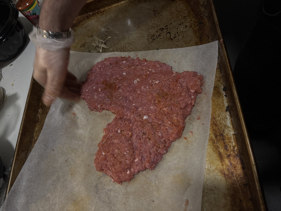 The image displays an overhead view of an indoor food preparation setting. A large, irregularly heart-shaped mass of raw, reddish-pink ground meat, approximately 15-20 cm at its widest point, is centered on a crinkled, off-white sheet of parchment paper. The meat exhibits flecks of lighter fat and dark seasoning granules. The parchment paper rests upon a tarnished, dark metallic baking sheet, which shows signs of use and heat discoloration.
A human hand, encased in a translucent white disposable glove, is positioned in the upper right quadrant, with fingers near the upper right curve of the meat. The hand appears blurred due to motion, indicating an active shaping or adjustment of the meat. A metallic wristwatch with a link band is visible on the gloved wrist.
In the blurred background, a dark pot or appliance and a partially visible, red-capped bottle are situated on a white countertop. Small, light-colored fibrous debris is present on the baking sheet to the right of the parchment paper. The scene is evenly and brightly lit. The image provides no visual indicators to determine its specific geographical location in the city of Sofia, Bulgaria.