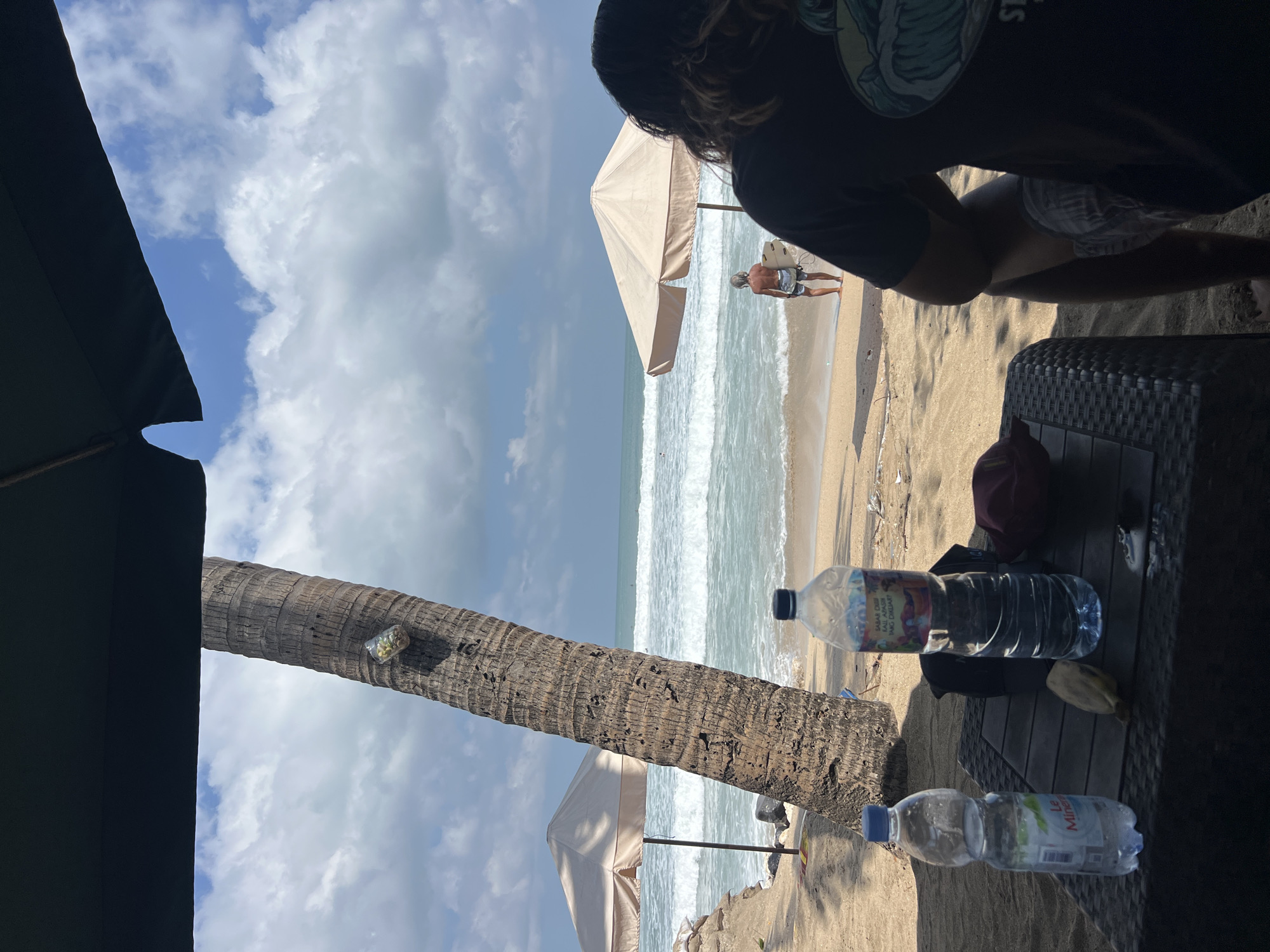 The image depicts a beach scene in Kuta, Indonesia, under a partly cloudy sky. In the foreground, a woven plastic table holds two plastic water bottles and a banana. A person with long, dark hair is seated at the table, facing away from the camera. In the middle ground, a palm tree stands to the left, with a plastic bag containing objects attached to its trunk. Two large beach umbrellas are visible, one on the left and another on the right. The ocean exhibits moderate waves, and a solitary surfer stands on the sand with a surfboard. The beach is sandy, with some debris scattered on the ground. The overall impression is a relaxed atmosphere at a popular Indonesian beach destination.