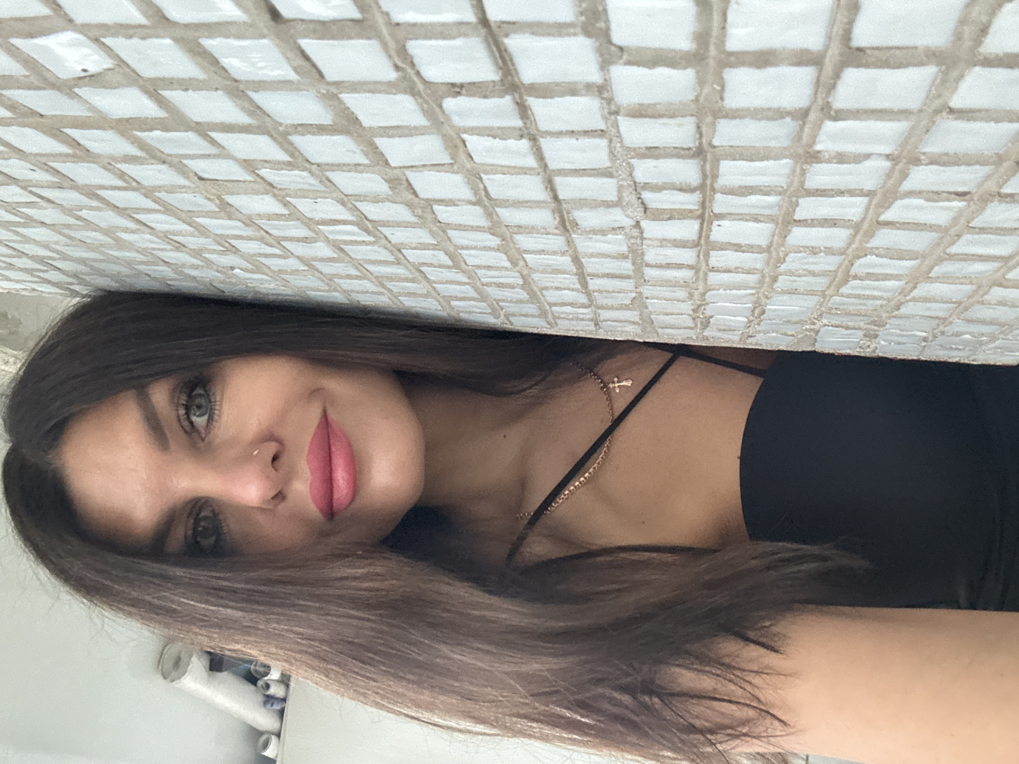 A close-up, waist-up photograph shows a woman with long, dark brown hair, fair skin, and green eyes. She is wearing a black top and a gold chain with a cross pendant. She is leaning against a tiled wall, which is light in color and has a grid pattern of small squares. The background on the left appears to be a shelf with some white cylindrical objects and other items. The image is taken from a slightly upward angle. The location is not specified in the image itself, and therefore cannot be determined to be in Chaikovsky, Russia.