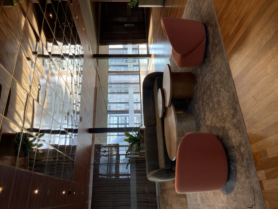 An interior scene depicts a mirrored wall on the left, reflecting light and patterns. The wall is adjacent to a tall window offering a glimpse of city buildings. A grey sofa and three round tables are situated on a patterned rug. Two coral-colored chairs flank the arrangement, one at each end. The right side of the image reveals a wooden floor, basking in sunlight. Plants are positioned near the window and the mirrored wall.