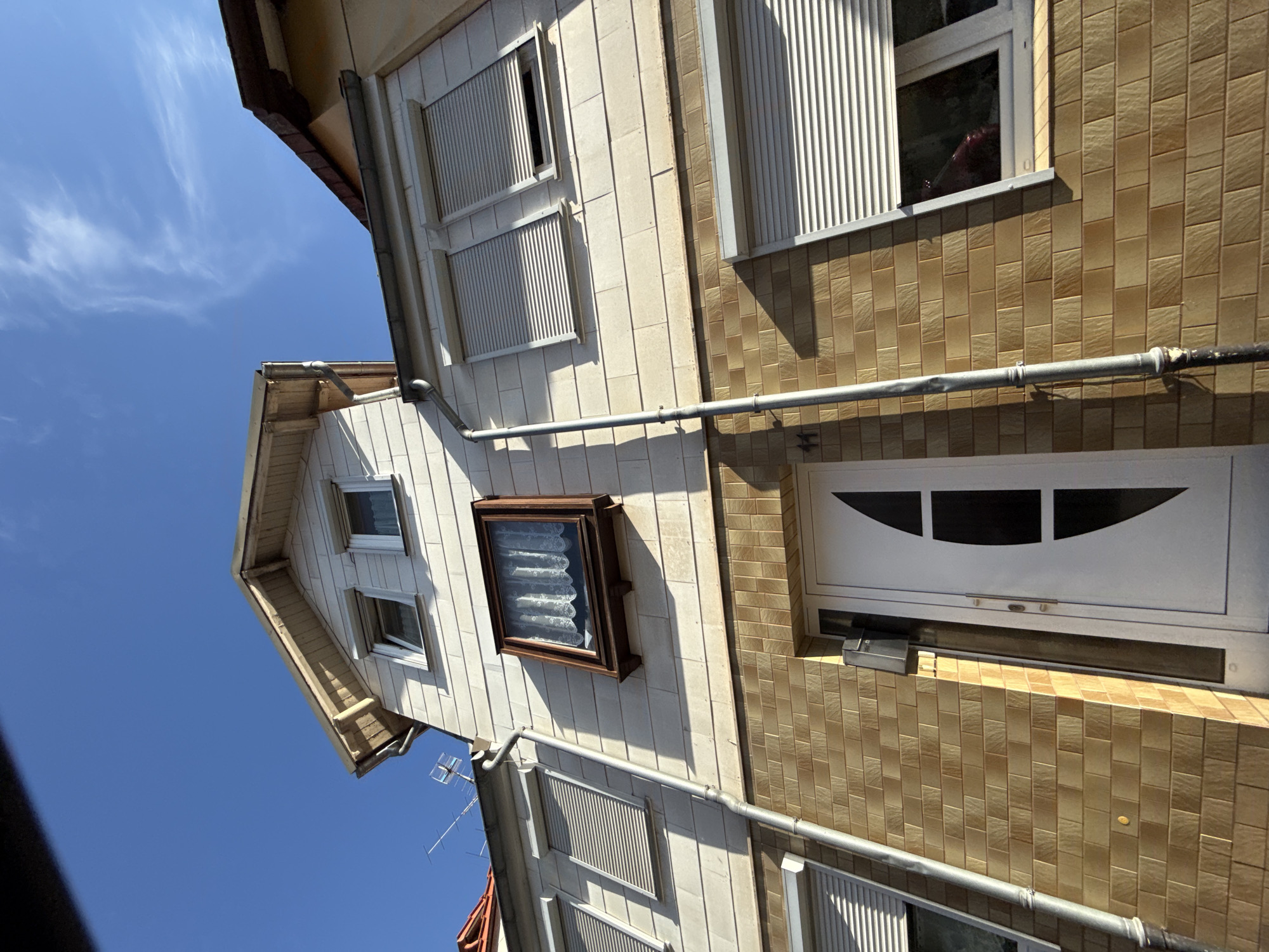 A low-angle, full-shot photograph captures the exterior of a light-colored, two-story house in Bad Sachsa, Germany. The facade of the lower story is clad in honey-colored brickwork, while the upper story is covered with white horizontal siding. A white front door with a dark, curved panel is visible on the lower story. Above the door, on the upper story, is a small, square window with white lace curtains. To the left of this window are two larger windows with closed, gray roller shutters. The roofline is visible at the top of the image, with exposed wooden beams. A drainpipe runs vertically down the side of the house, crossing both stories. The sky above is a clear, bright blue with a few wispy white clouds. The sun casts strong shadows, indicating it is daytime.