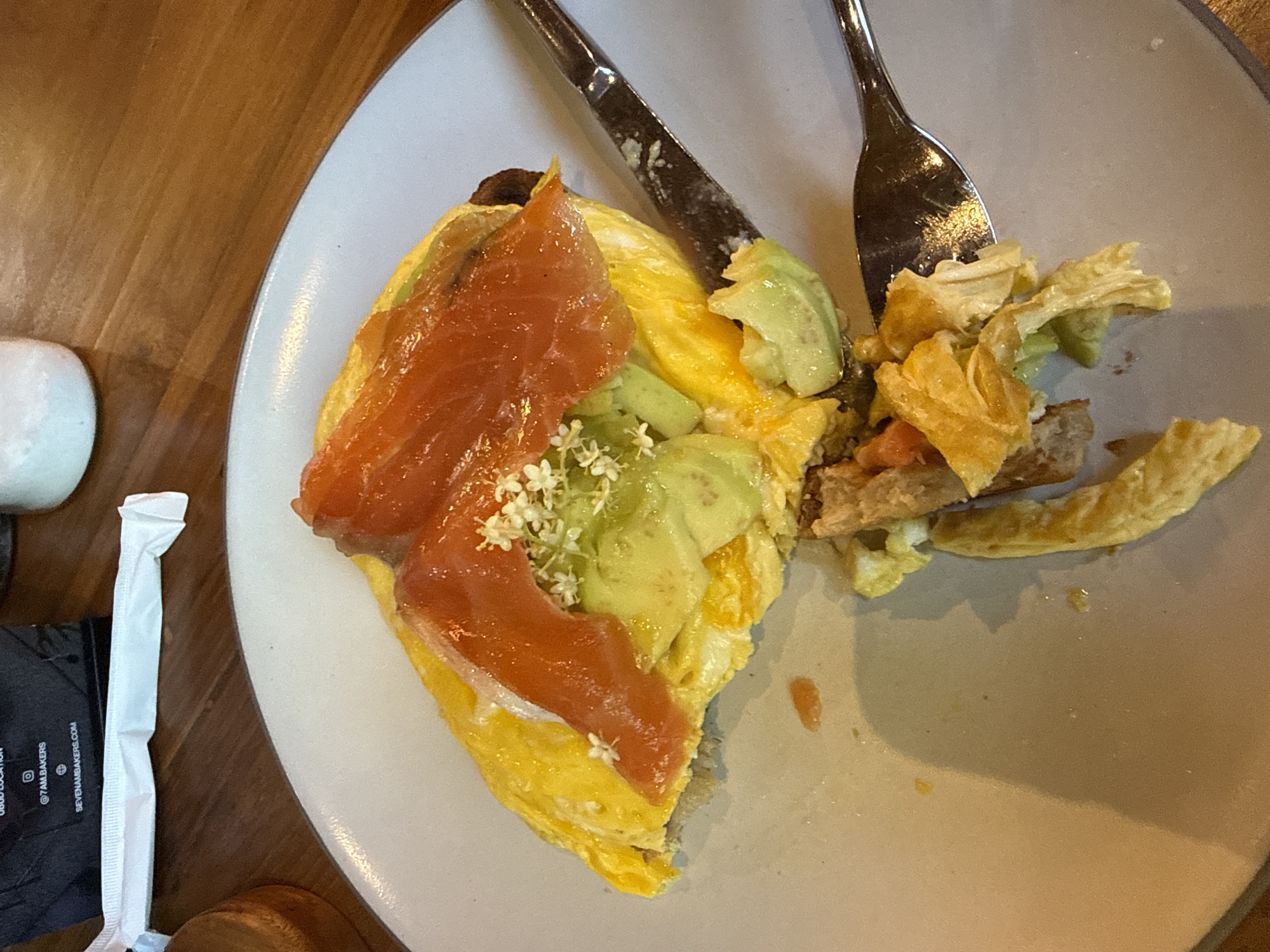 A high-angle shot shows a partly eaten meal on a light grey plate, likely in a restaurant setting, though Ubud District, Indonesia, is not visibly confirmed. The meal consists of an omelet topped with salmon, avocado, and small white flowers. A piece of bread is also visible. A fork and knife rest on the plate, indicating the meal is partially consumed. Next to the plate, a small, white, wrapped object, likely a utensil, and a black card with white writing are visible. The plate sits on a wooden surface.