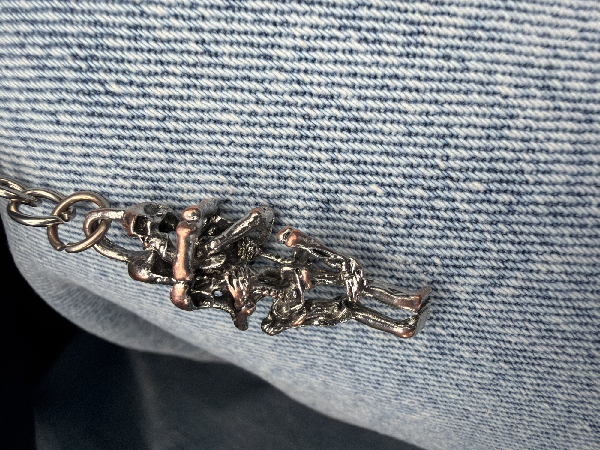 A close-up photograph captures a metallic keyring adorned with several intricate charms, set against a light blue denim fabric background. The charms are clusters of small, highly detailed skeletal figures, appearing to be human skeletons or similar bone structures. These figures display a dual-tone metallic finish, with certain parts, such as bones or joints, exhibiting a reddish-bronze or copper hue, contrasting with the darker, possibly oxidized silver or gunmetal finish of other sections. The individual skeletal elements are interconnected, forming a dense and complex assemblage of figures. The keyring component, visible on the left, is a bright silver-colored metal.
The background is dominated by the textured weave of the denim fabric, which features prominent diagonal lines of blue and white threads characteristic of its twill construction. The denim appears slightly worn or faded in areas, accentuating its tactile quality. A darker, blurred region occupies the lower left of the frame, possibly indicating a shadow or another part of the garment out of focus. The lighting is soft and even, highlighting the metallic sheen of the charms and the texture of the fabric. No activity or event is depicted, nor are there any people, visible text, or specific location cues beyond the denim material itself.