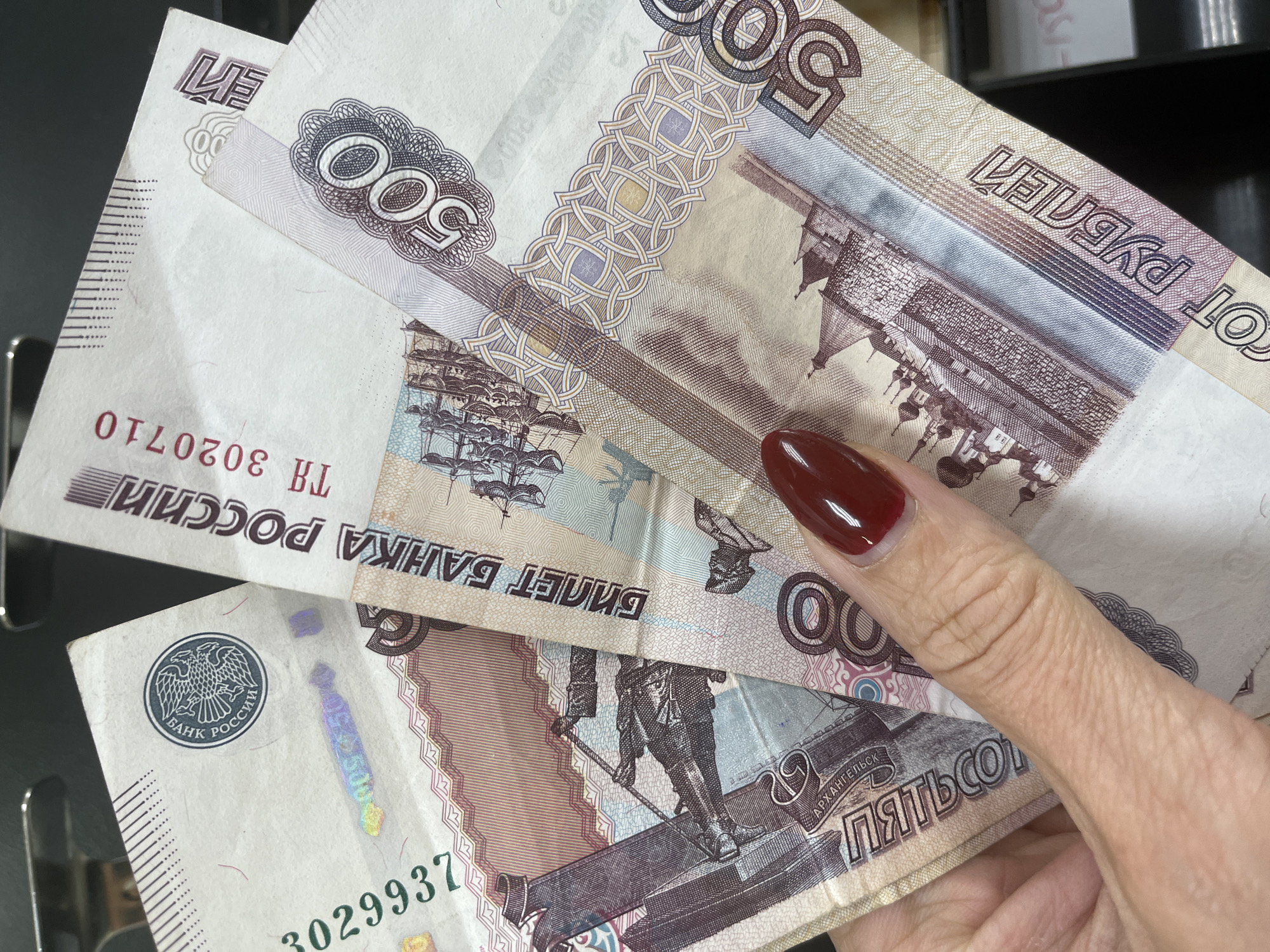 A close-up, top-down, eye-level shot shows a person's hand holding several Russian banknotes. The banknotes are partially fanned out, with their serial numbers and denominations visible. The hand has a dark red nail polish on the index fingernail. The background is dark and out of focus, suggesting an indoor setting. The banknotes appear to be of denominations 500 rubles and 100 rubles. One note shows the text "БАНК РОССИИ" (Bank of Russia) and a two-headed eagle emblem. Another note shows the number "500" prominently. A third note shows the number "100" and the text "ПЯТЬСО" (Five hundred) and "АРХАНГЕЛЬСК" (Arkhangelsk), suggesting it is a 500 ruble note depicting Arkhangelsk. Another note visible has the serial number "3029937" and possibly "TH 3020710". The banknotes are in good condition.