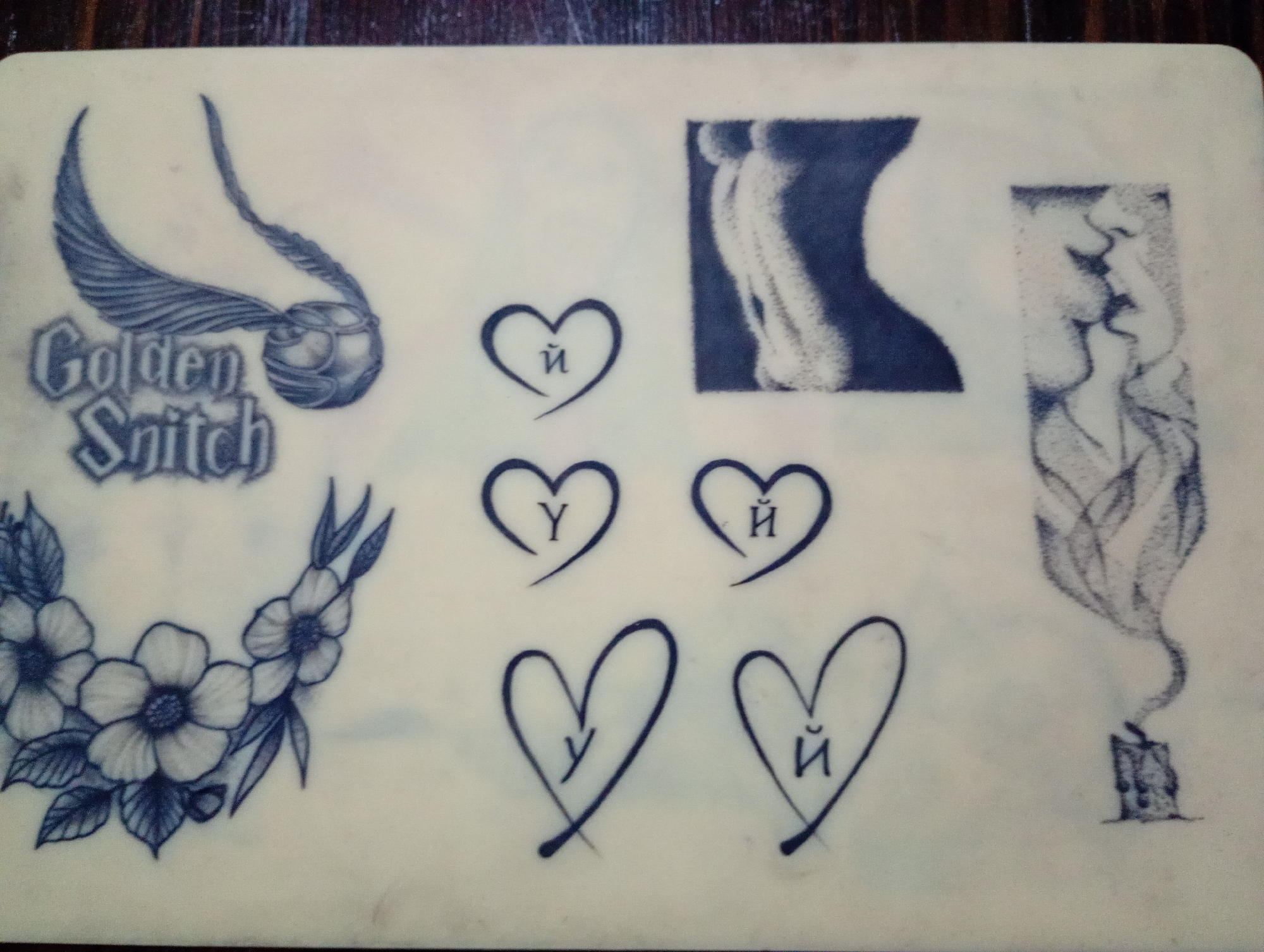 The image shows a tattoo practice skin, likely made of silicone or a similar material, with various tattoo designs applied in dark blue ink. On the left, there is a rendering of a Golden Snitch from the Harry Potter series, along with the text "Golden Snitch," and a floral design featuring flowers and leaves. The center shows four heart designs, each containing either the Cyrillic letter "У" (U) or "Й" (short I). To the right of these heart designs are two shaded square images. The first appears to be an abstract representation of a body. The second square image shows two faces in profile in the midst of kissing. To the far right is a candle illustration in the midst of burning. There is no information in this image to ascertain its location in Roza, Bulgaria, or any city for that matter.