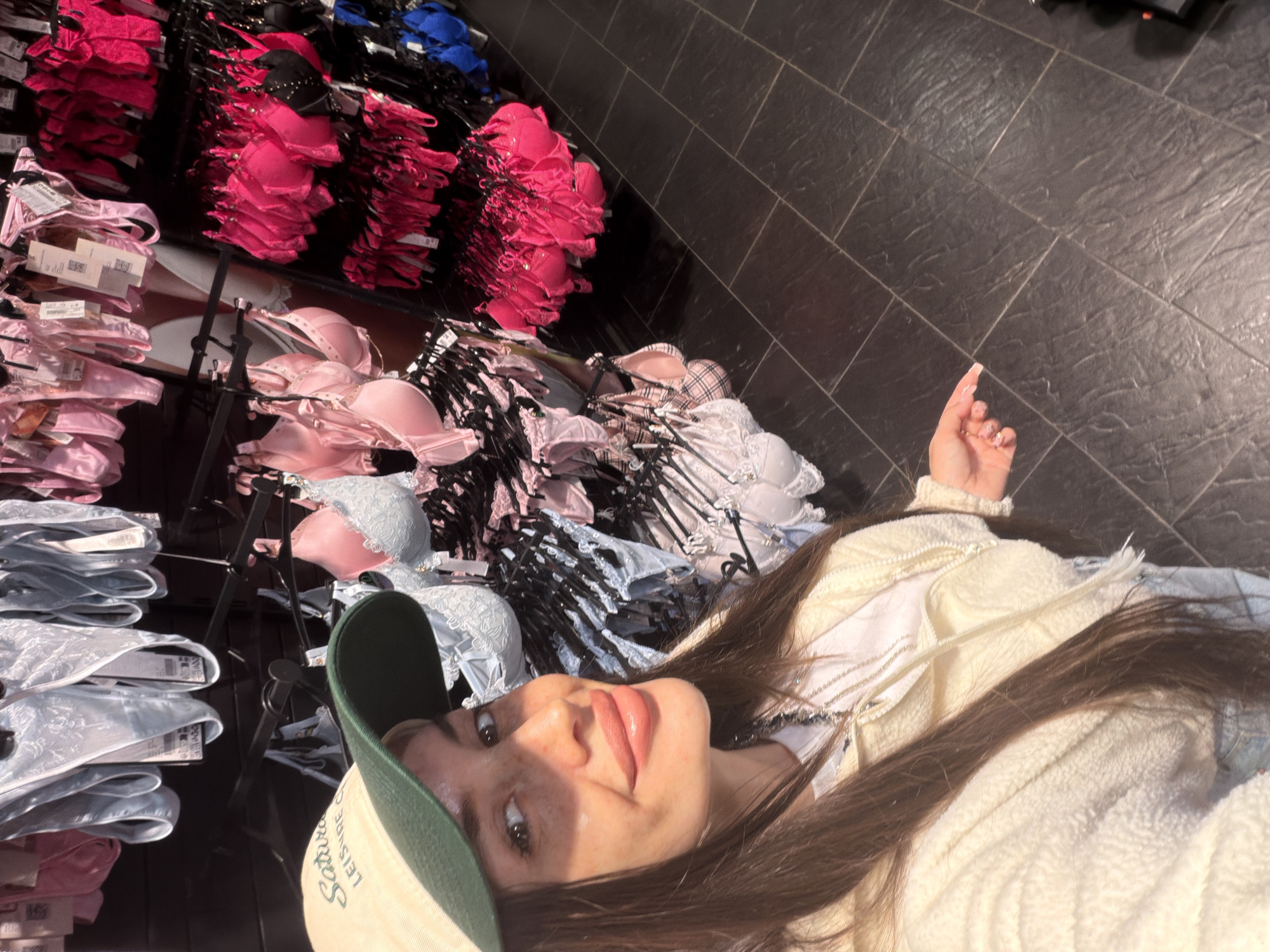 A woman wearing a cream-colored fuzzy jacket and a tan baseball cap with green accents is in a store, possibly a lingerie shop. She is looking at the camera and smiling. Behind her, racks of lingerie are displayed, including bras and underwear in various colors such as pink, blue, and white. The floor is tiled in a dark color. The scene is indoors, and there is no indication of its specific location in Saint Petersburg, Russia.