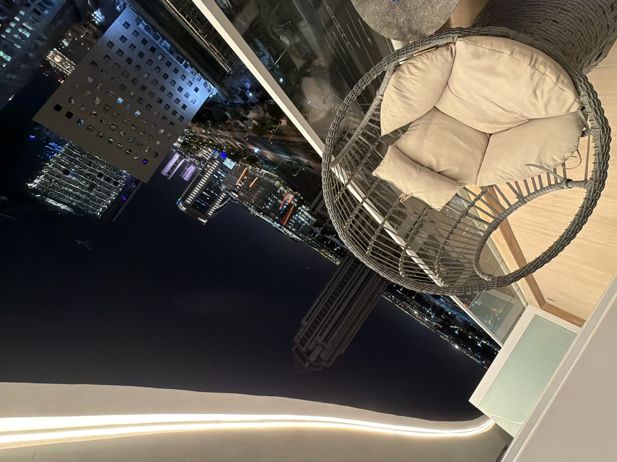 The image is a tilted, high-angle shot of a balcony at night, overlooking a city skyline. In the foreground, a grey, wicker hanging chair with light brown cushions is prominently displayed. The chair is empty and appears to be outdoors, likely on a balcony. Beyond the chair, a glass railing separates the balcony from the city view.
The city skyline features numerous modern skyscrapers illuminated with bright lights. The buildings vary in height and architectural style, with some displaying intricate patterns of windows and lights. Roads with moving car lights are visible between the buildings, indicating a busy urban environment. The sky is dark, suggesting it is nighttime.
To the left of the image, a portion of a white, curved architectural element is visible, possibly part of the building's exterior or a decorative lighting fixture. This element is illuminated with a soft, warm light.
The overall scene conveys a sense of urban living and nighttime ambiance. The orientation of the camera suggests the photographer was looking down and to the side from a higher vantage point.