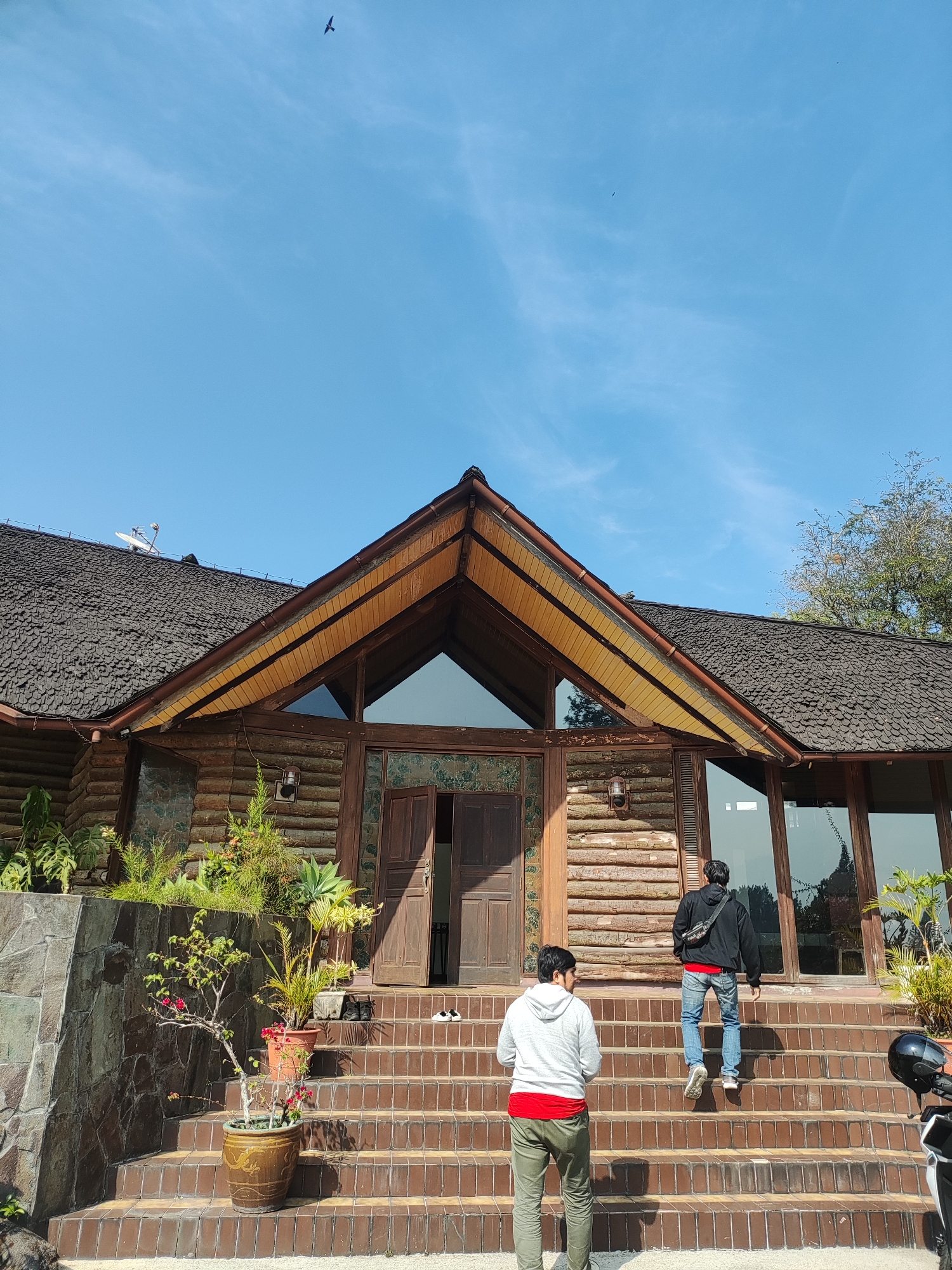 In Cisarua, Indonesia, a wide shot captures a log cabin-style building with a prominent A-frame entrance. Two young men are walking up a wide, tiled staircase leading to the entrance. The man in the foreground, wearing a grey hoodie over a red shirt and green pants, faces away from the camera. The man behind him, dressed in a black hoodie, blue jeans, and a red shirt visible at the waist, walks up the stairs. The building's exterior features wooden log construction and a textured, dark roof. Lush greenery is visible around the base of the building and on the stairs. A satellite dish is mounted on the roof. The sky is clear blue with a few wispy clouds and a single bird in flight.
