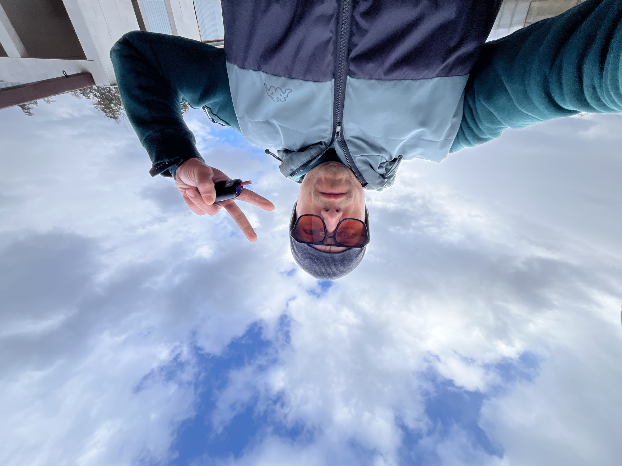 The image presents a man in an inverted orientation, appearing upside down, with a cloudy sky dominating the background. He is looking directly towards the camera.
The man wears a two-tone puffer jacket, dark blue or black on the shoulders and upper chest, transitioning to a lighter blue or grey on the lower half. A dark teal or green long-sleeved shirt is visible underneath. He is also wearing a grey beanie and dark sunglasses with reddish-brown lenses, which obscure his eyes. A small white logo, resembling the Kappa brand's twin silhouette, is visible on the light blue section of his jacket.
In his left hand, he holds a small, dark cylindrical object, possibly a vape pen, and simultaneously makes a peace sign with his index and middle fingers. A dark smartwatch is worn on his left wrist.
The setting is outdoors, characterized by a bright, overcast sky with areas of clear blue visible through the clouds, indicating daytime. In the upper portion of the inverted image (which would be at the bottom if correctly oriented), structural elements of what appears to be a building's roof or an overhang are visible. Some blurred, bare tree branches can also be seen on the left side.
The activity appears to be a selfie or a photo taken from a very low angle, resulting in the unusual upside-down perspective. The man's pose and the peace sign suggest a casual or relaxed mood. The image provides no specific visual cues to indicate the given location context of Chaikovsky, Russia.