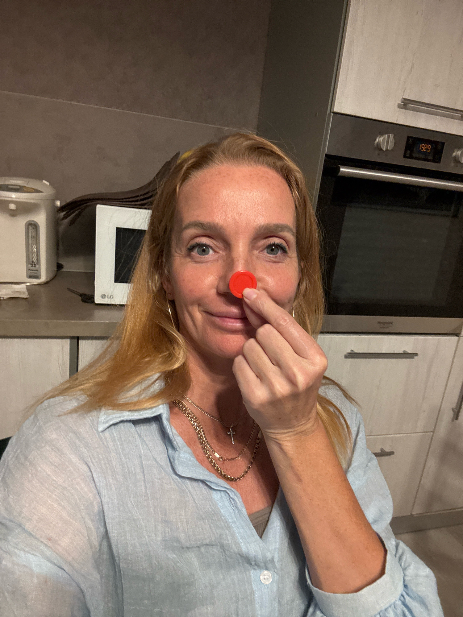 A blonde woman with fair skin and freckles is standing in a kitchen. She is wearing a light blue collared shirt and several necklaces. She is holding a small red object on her nose with her right index finger and thumb. In the background, there is a microwave, an oven, and kitchen cabinets. The lighting is artificial and casts shadows. The image appears to be a selfie taken by the woman. The location is not specified in the image or metadata.