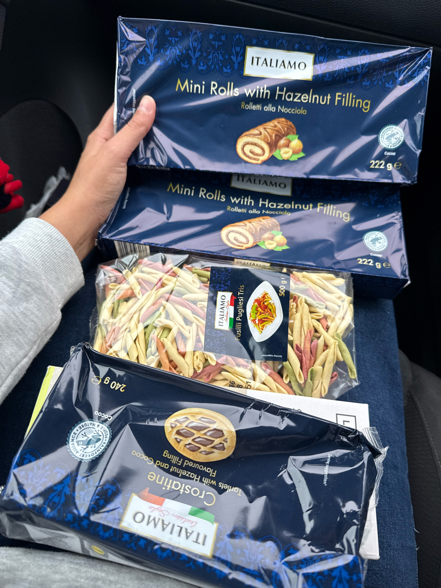 The image shows a person's hand holding several packages of Italianmo brand food products. The packages are stacked vertically. The top two packages are dark blue boxes labeled "ITALIAMO Mini Rolls with Hazelnut Filling". Below these are two plastic-wrapped packages of pasta. The upper pasta package contains tri-colored fusilli. The bottom package is a dark blue wrapper with a picture of a pastry, labeled "ITALIAMO Crostatine". The scene appears to be inside a vehicle, possibly a car, as suggested by the dark interior and seat fabric. No specific landmarks of Great Yarmouth are visible.