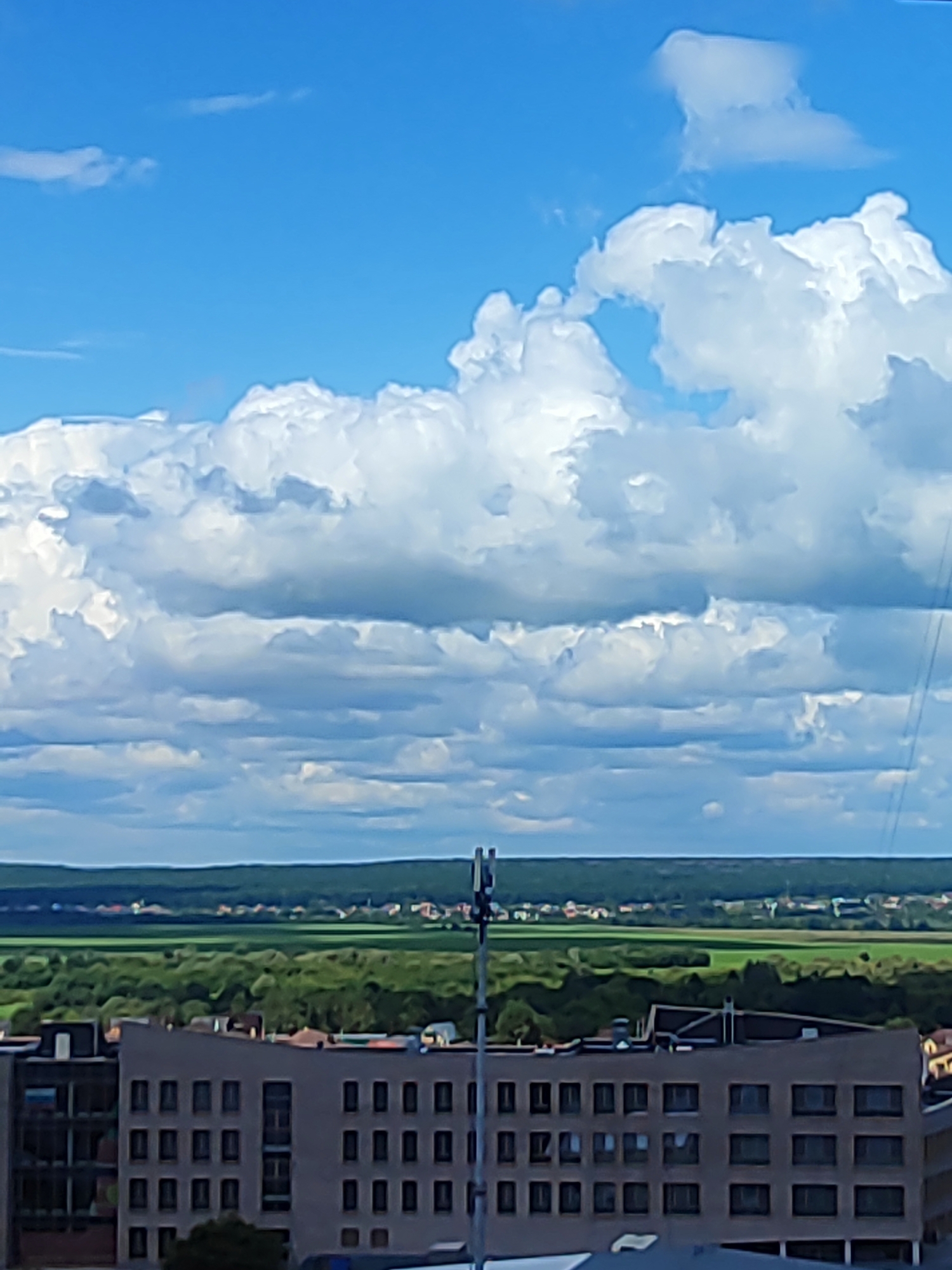 The image captures a wide shot of a cityscape on a cloudy day. In the foreground, a modern, multi-story building with a dark facade and numerous windows dominates the lower portion of the frame. A tall cellular tower, topped with antennas, stands to the right of the building. Beyond the building, a vast expanse of green fields and trees stretches to the horizon, dotted with small houses and structures. The sky above is a vibrant blue, filled with dramatic, voluminous white and gray clouds. The scene appears to be an urban or semi-urban area, likely a suburb or outskirts, given the mix of buildings and natural landscapes. The overall atmosphere is bright but overcast.