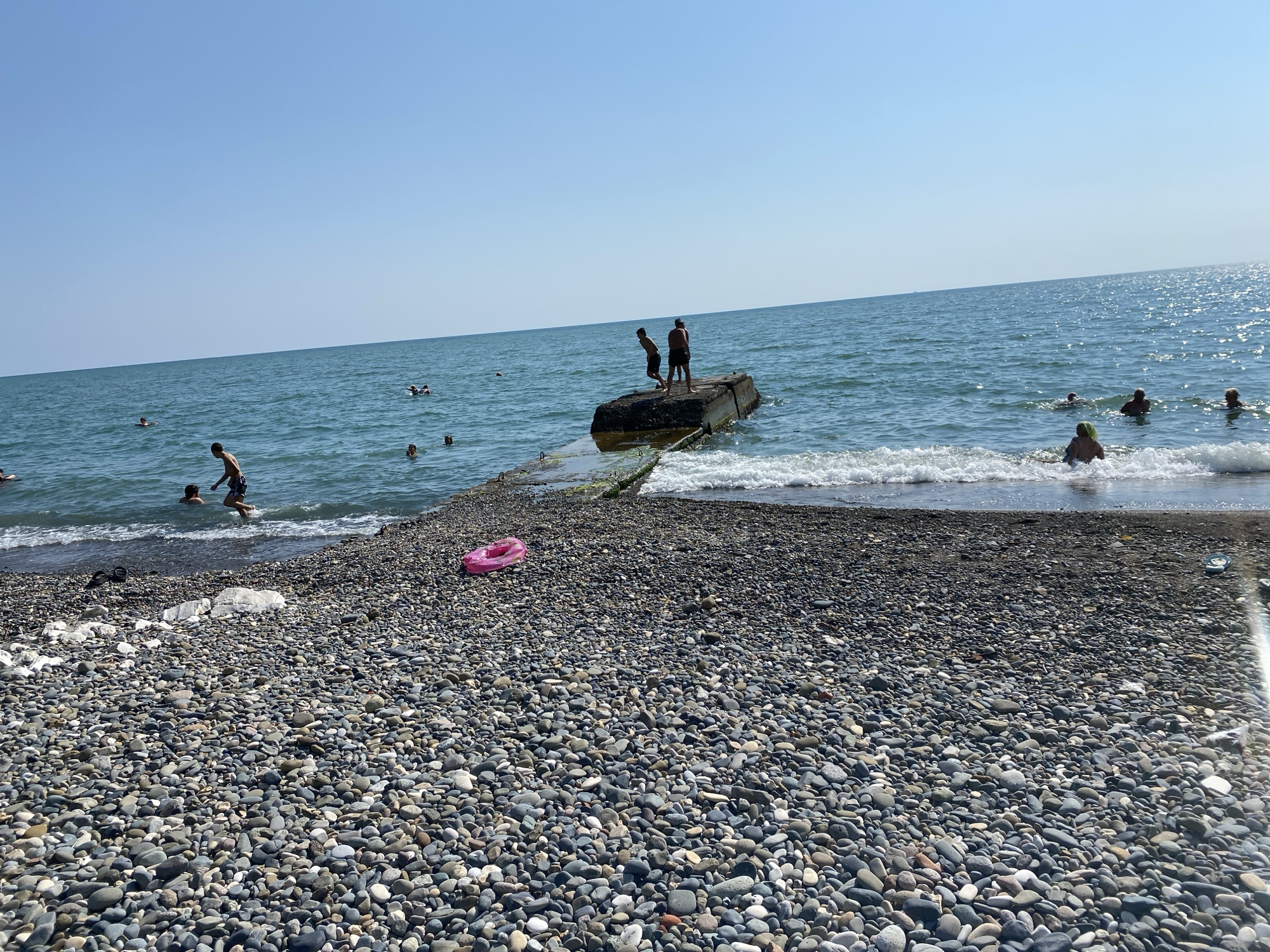 In Chabkwara, Abkhazia, a sunny day at a pebble beach is depicted. The foreground is dominated by a vast expanse of grey and colored pebbles. The turquoise sea stretches to the horizon under a clear blue sky. Several people are visible in the water, some swimming and others wading near the shore. In the middle ground, two young males are standing on a concrete structure extending into the sea, with one appearing to be jumping into the water. Further out, more individuals are enjoying the sea. A bright pink inflatable ring lies on the pebbles near the water's edge. The scene captures a typical summer day at the beach.