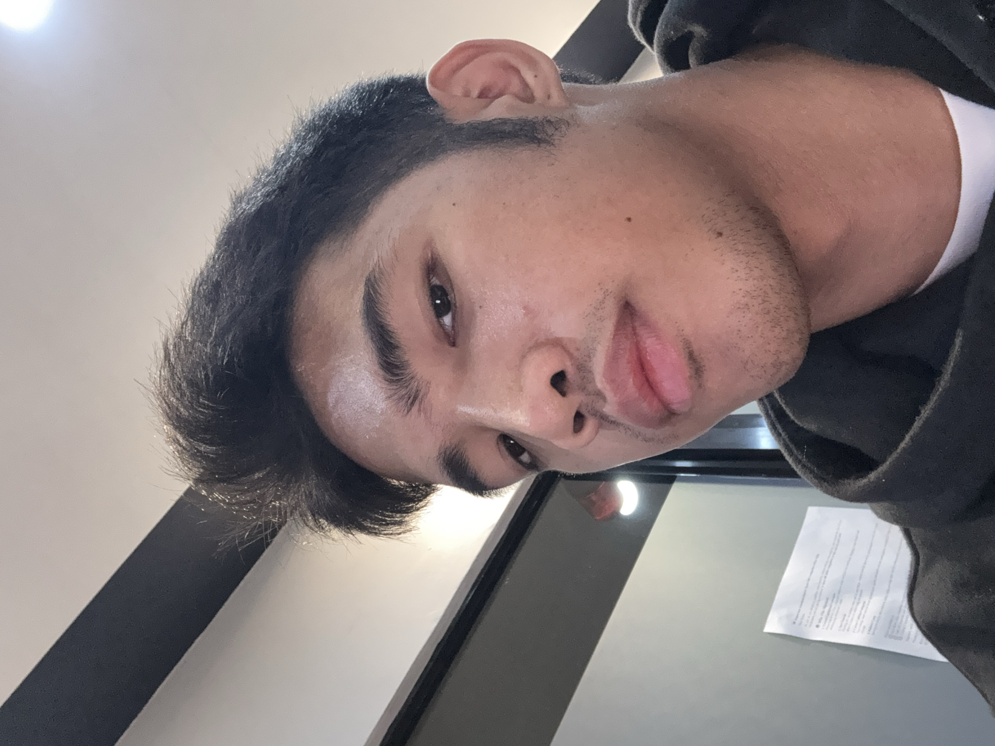 The image is a close-up, low-angle selfie of a young man with dark hair styled upwards and a faint mustache and stubble. He is wearing a black hooded sweatshirt over a white t-shirt. The background appears to be an indoor environment with a gray wall on the left, a white ceiling with a dark beam, and what looks like a document on the wall. There are also bright lights visible in the background. The lighting suggests it is daytime or a well-lit interior. The image's composition and focus are on the man's face, with his expression neutral and his gaze directed slightly off-camera. No specific landmarks or recognizable features of Davao City are visible in this cropped image.