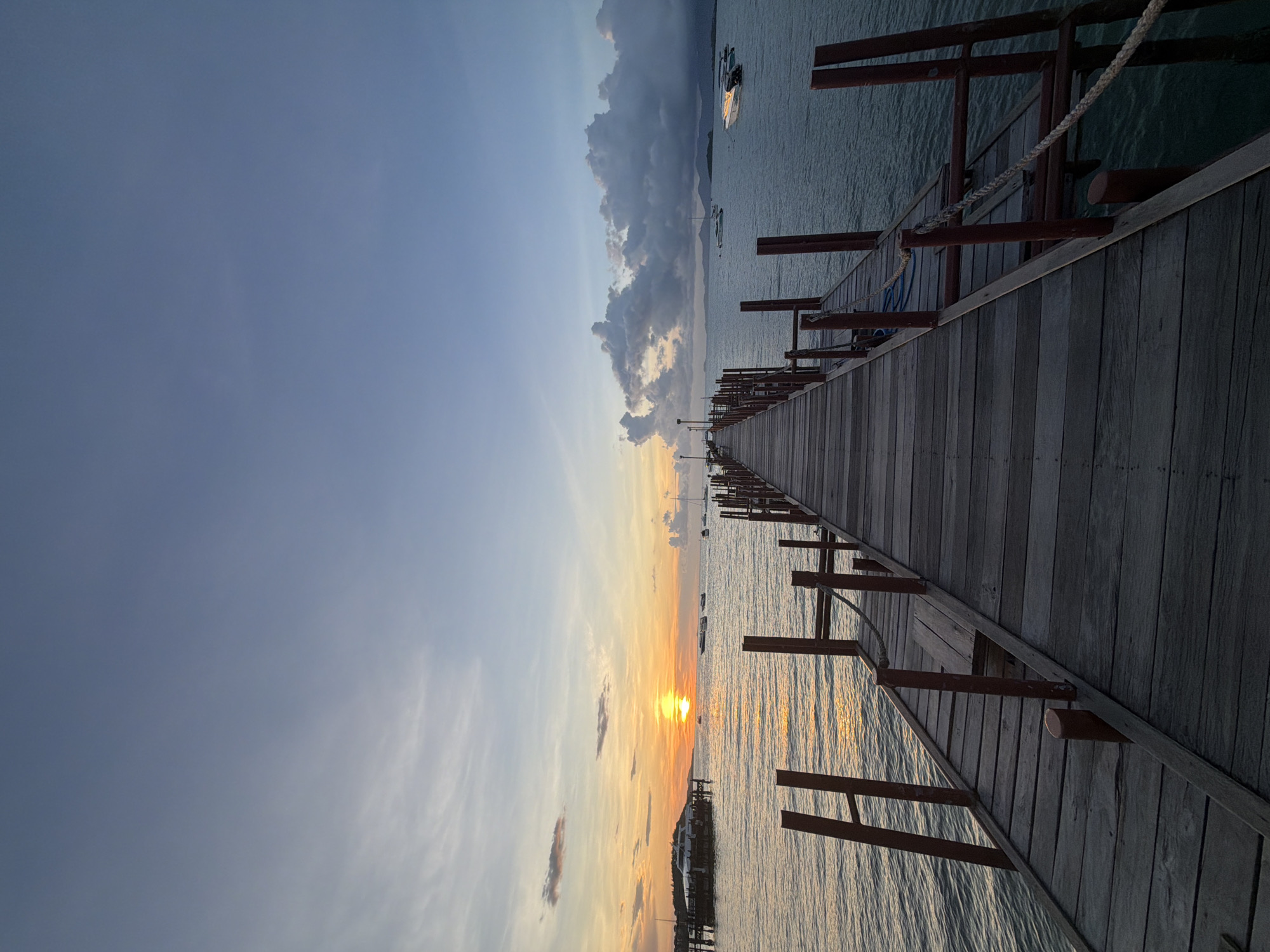 A wide-angle, vertical shot captures a wooden pier extending into the ocean at sunset in Baan Bang Rak, Thailand. The sun, a bright orange orb, is setting over the water on the left side of the frame, casting a golden reflection across the gentle waves. Wispy clouds streak across the sky, transitioning from warm oranges and yellows near the horizon to a pale blue overhead. Several boats, including a ferry and smaller vessels, are visible in the distance. The pier itself is constructed of weathered wooden planks and is lined with reddish-brown wooden posts, some with ropes attached. The water is a deep blue, with a slight rippling texture. The overall atmosphere is serene and peaceful, with a sense of natural beauty and tranquility.