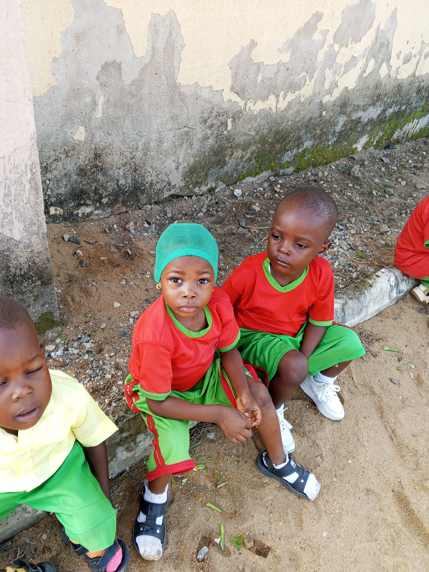 Four young children are seated on the sandy ground in front of a weathered, light-colored wall in Akwanga, Nigeria. The wall exhibits patches of peeling paint and discoloration, with streaks of moss and lichen. Two of the children, a boy and a girl, are wearing matching red t-shirts with green trim and green shorts. The girl has a green headscarf. The other two children, both boys, are dressed in a light yellow collared shirt and green pants, and another boy is partially visible in a red shirt. The ground is composed of sand and small pebbles, with some dried leaves and twigs scattered about. The overall impression is of children taking a break outdoors, possibly at a school or community gathering.