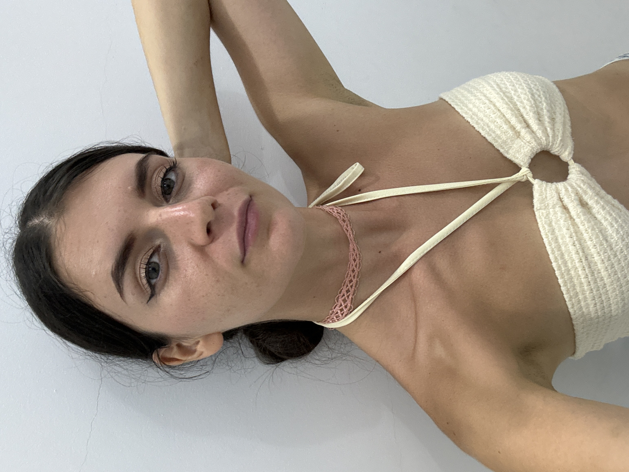 A woman with dark hair and fair skin is lying on a white surface. She is wearing a white crocheted top with straps that tie around her neck, where she is also wearing a light pink crocheted choker. Her arms are raised above her head. The image appears to be taken indoors with natural lighting. The location is not discernible from the provided image.